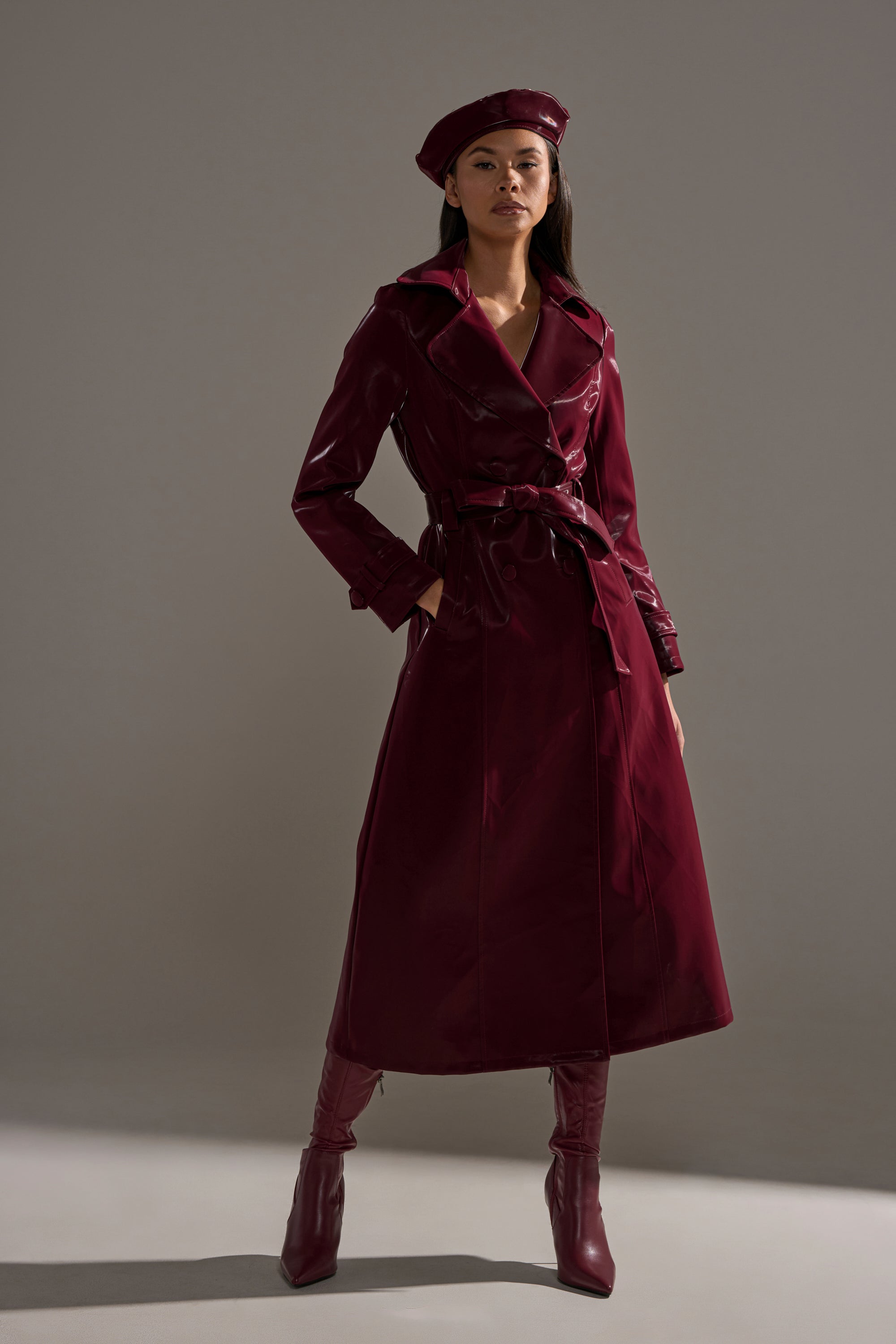A woman stands confidently against a plain gray background wearing the SANDO LIQUID VELVET TRENCH IN BURGUNDY, paired with matching knee-high boots and a beret.
