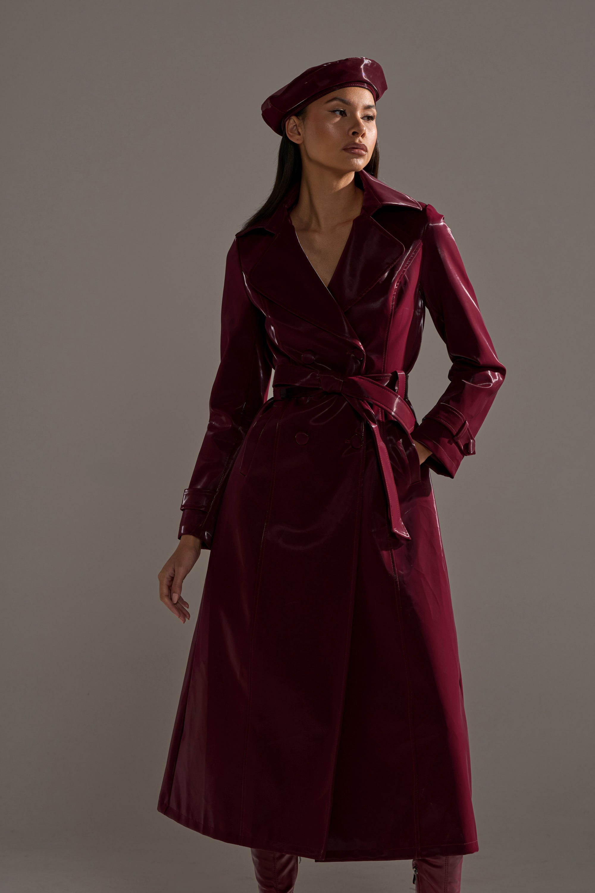 A woman poses in the SANDO LIQUID VELVET TRENCH IN BURGUNDY with a matching belt and beret, looking to the side against a plain gray background.