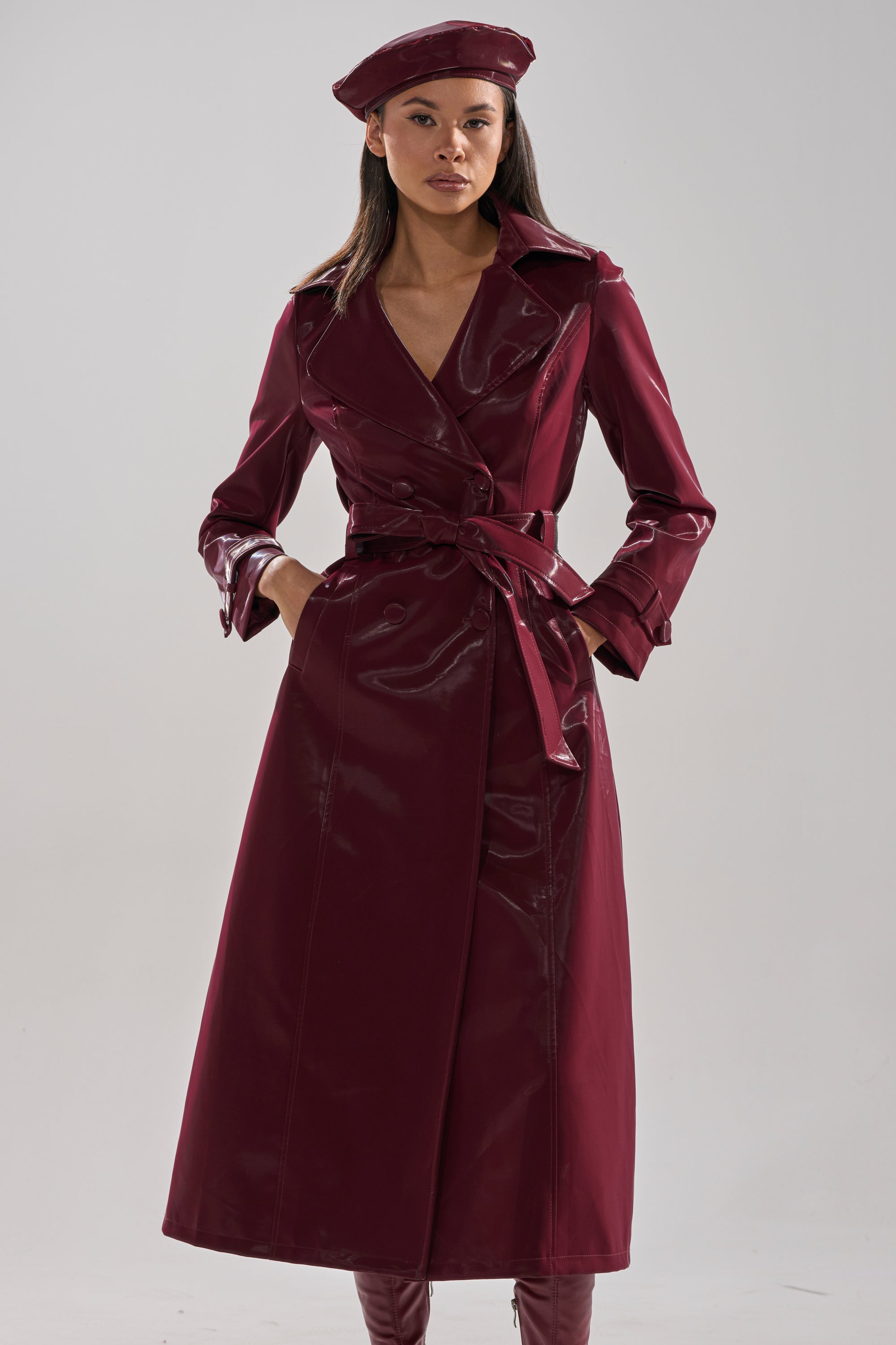 A woman stands confidently against a plain background, wearing the AKIRA Label SANDO LIQUID VELVET TRENCH IN BURGUNDY with a matching beret and tall boots, looking directly at the camera.