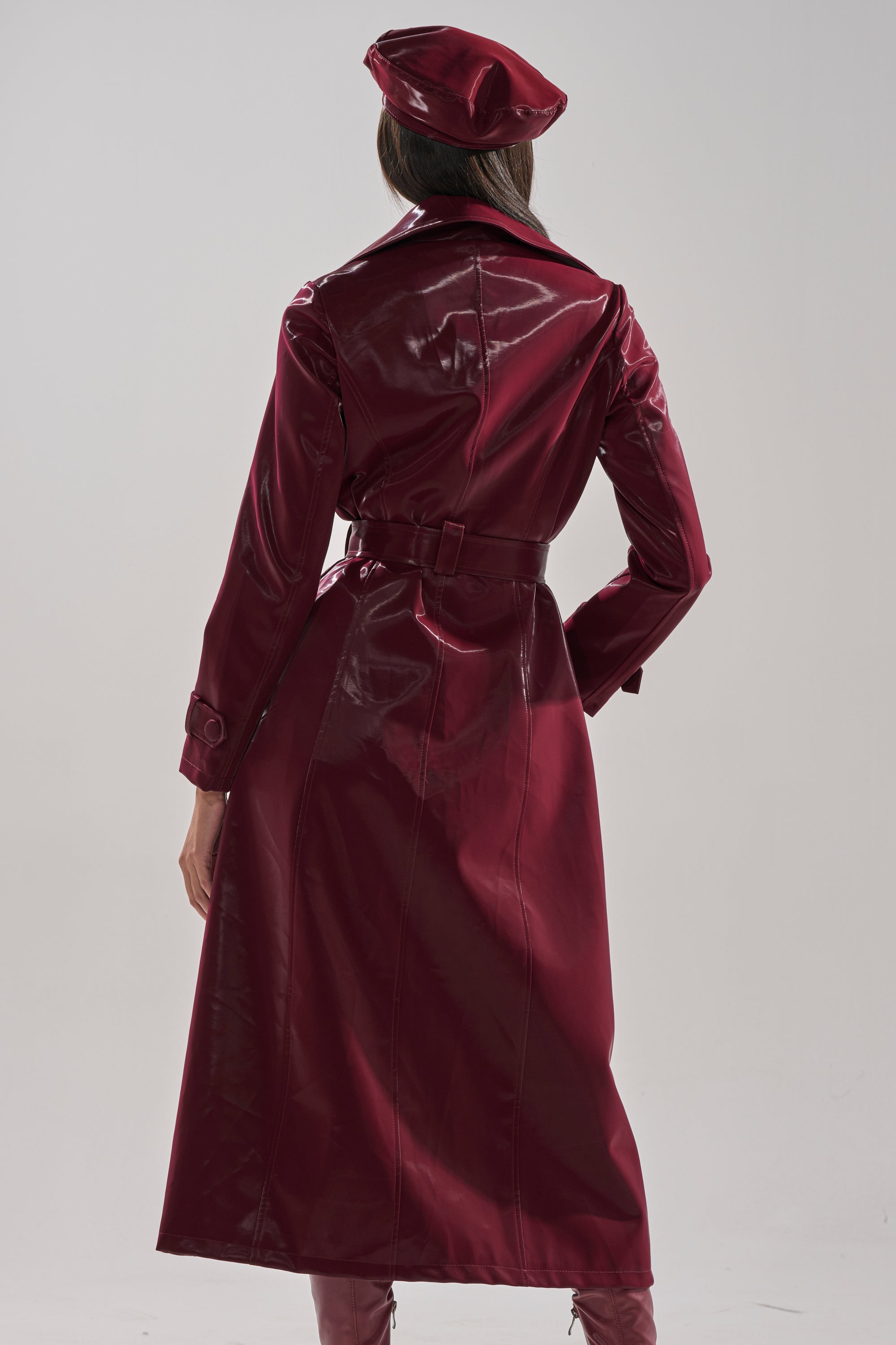 A person faces away, wearing the SANDO LIQUID VELVET TRENCH IN BURGUNDY—a glossy, faux leather coat with matching belt and beret from AKIRA Label. The outfit has a sleek, reflective finish against a plain, light background.