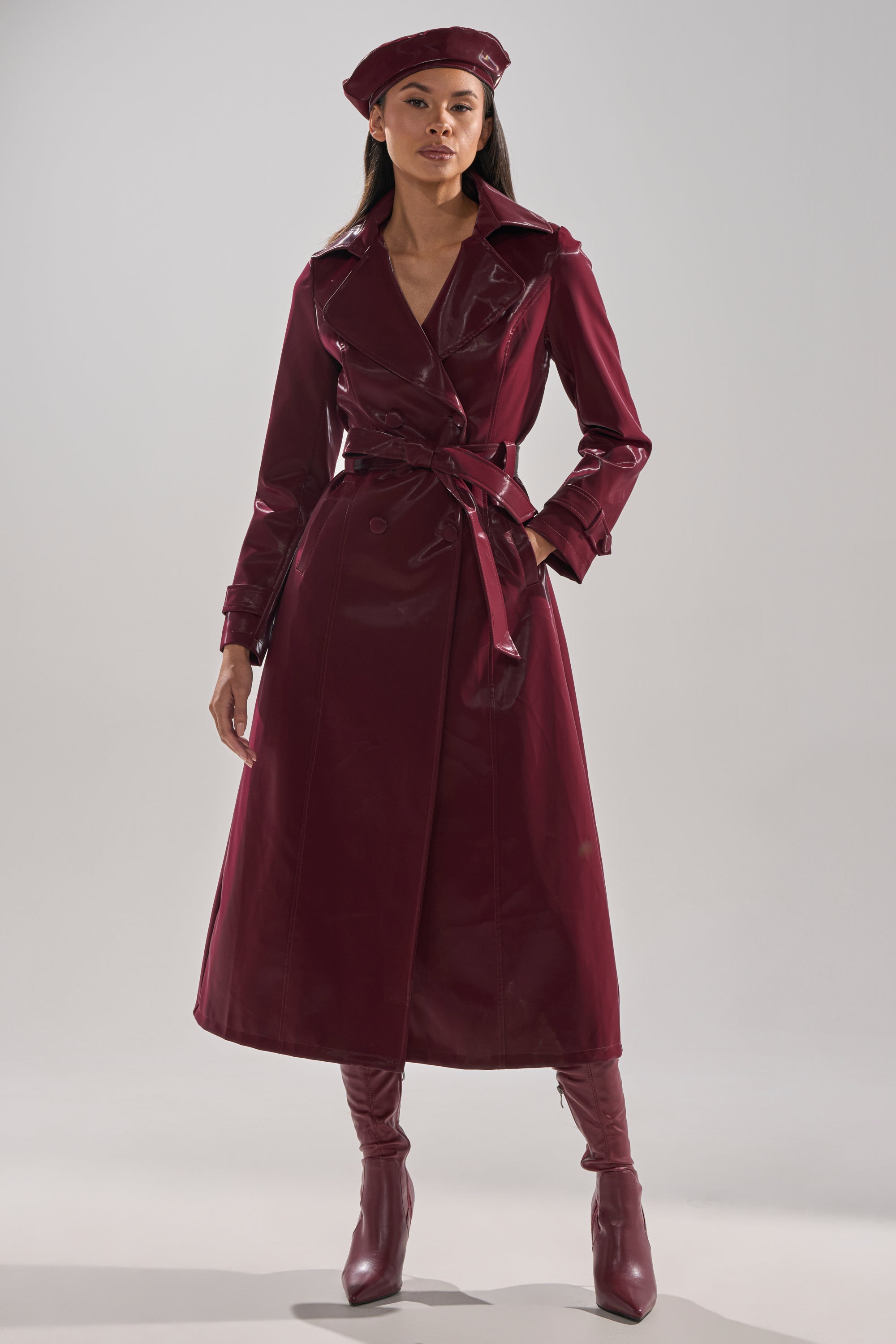 A woman poses confidently against a plain light background in the SANDO LIQUID VELVET TRENCH IN BURGUNDY, with matching knee-high boots and a beret.