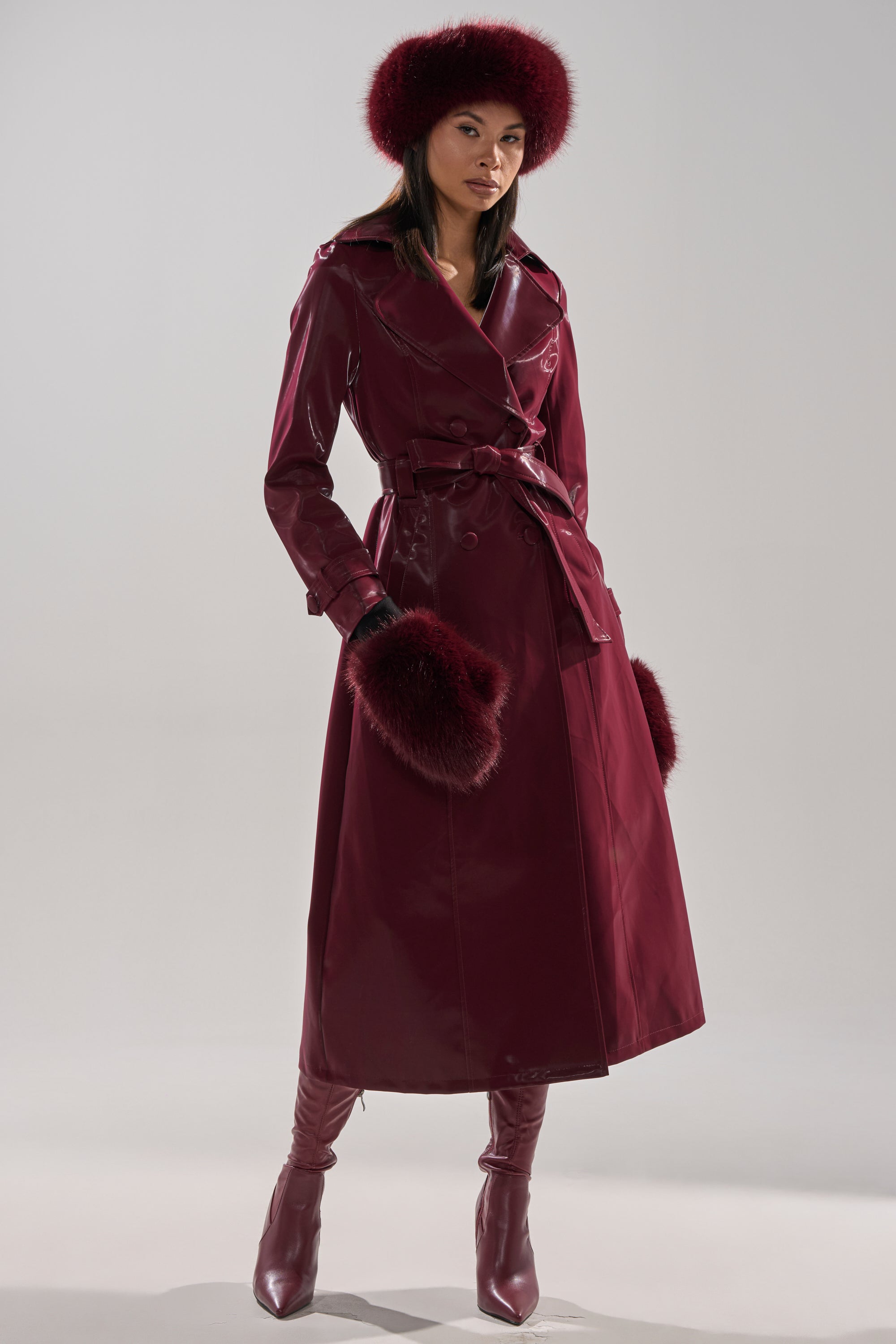 A woman models the SANDO LIQUID VELVET TRENCH IN BURGUNDY, featuring a matching belt, fur-lined hat, fur cuffs, and knee-high boots against a minimalist light background.