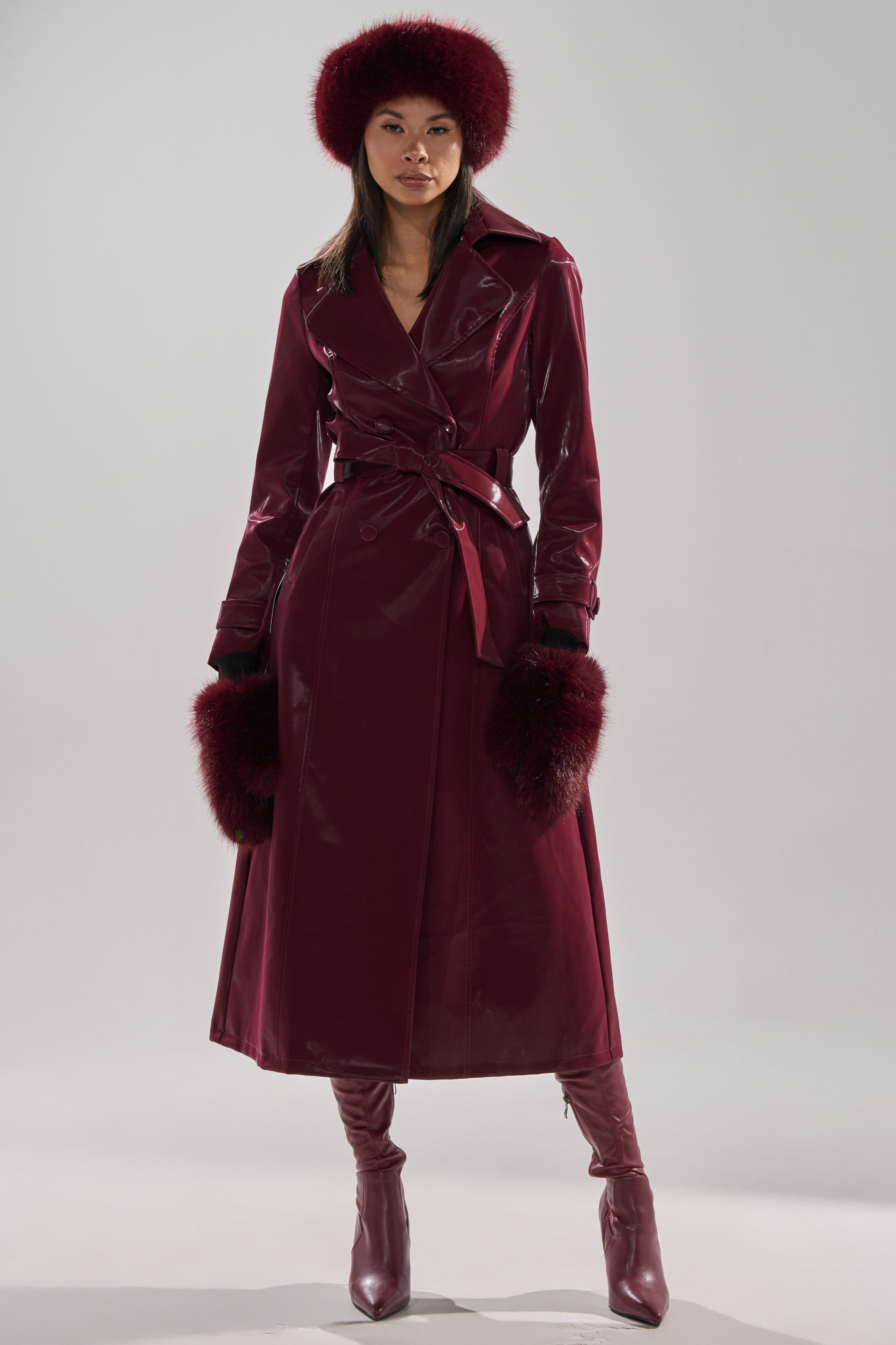 A woman exudes confidence in the SANDO LIQUID VELVET TRENCH IN BURGUNDY, paired with tall boots, a matching fur hat, and fur-trimmed gloves, against a plain light backdrop—showcasing modern elegance in rich burgundy tones.