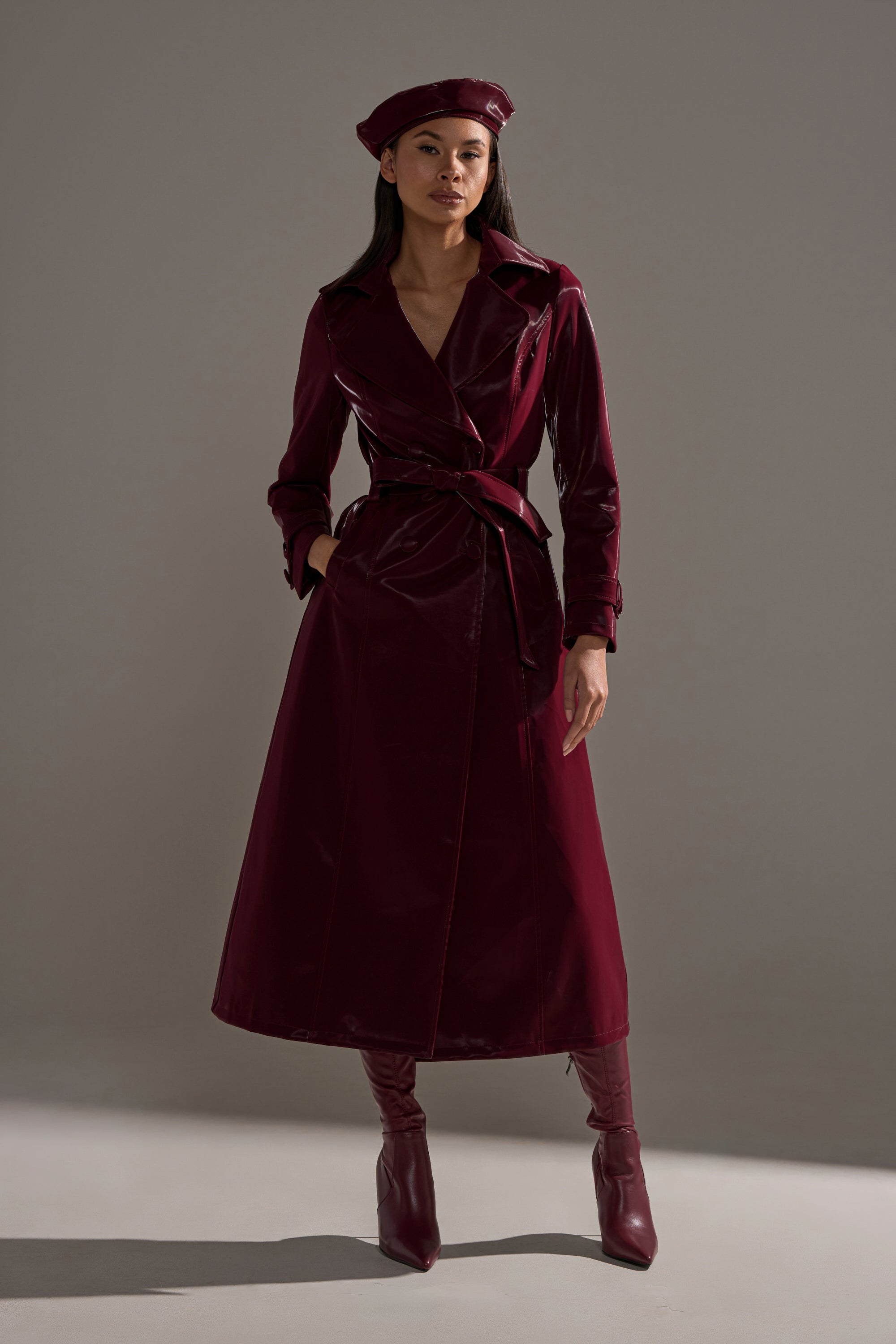 A woman stands confidently with hands in pockets, wearing the SANDO LIQUID VELVET TRENCH IN BURGUNDY, belted at the waist and paired with matching knee-high boots and a beret, set against a plain gray background.