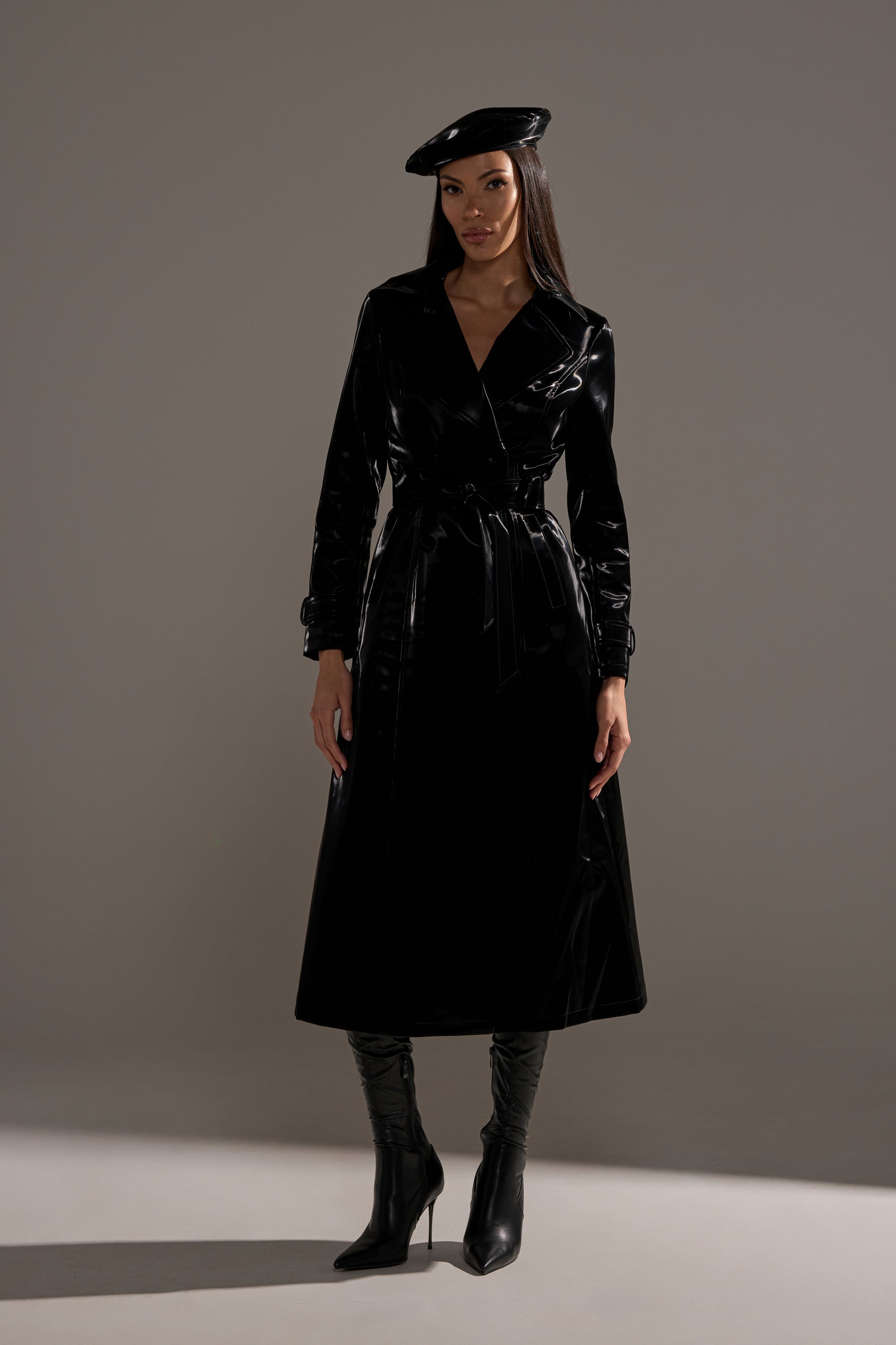 A woman poses in a studio against a gray backdrop, wearing the SANDO LIQUID VELVET TRENCH IN BLACK paired with a black beret and knee-high black heeled boots.