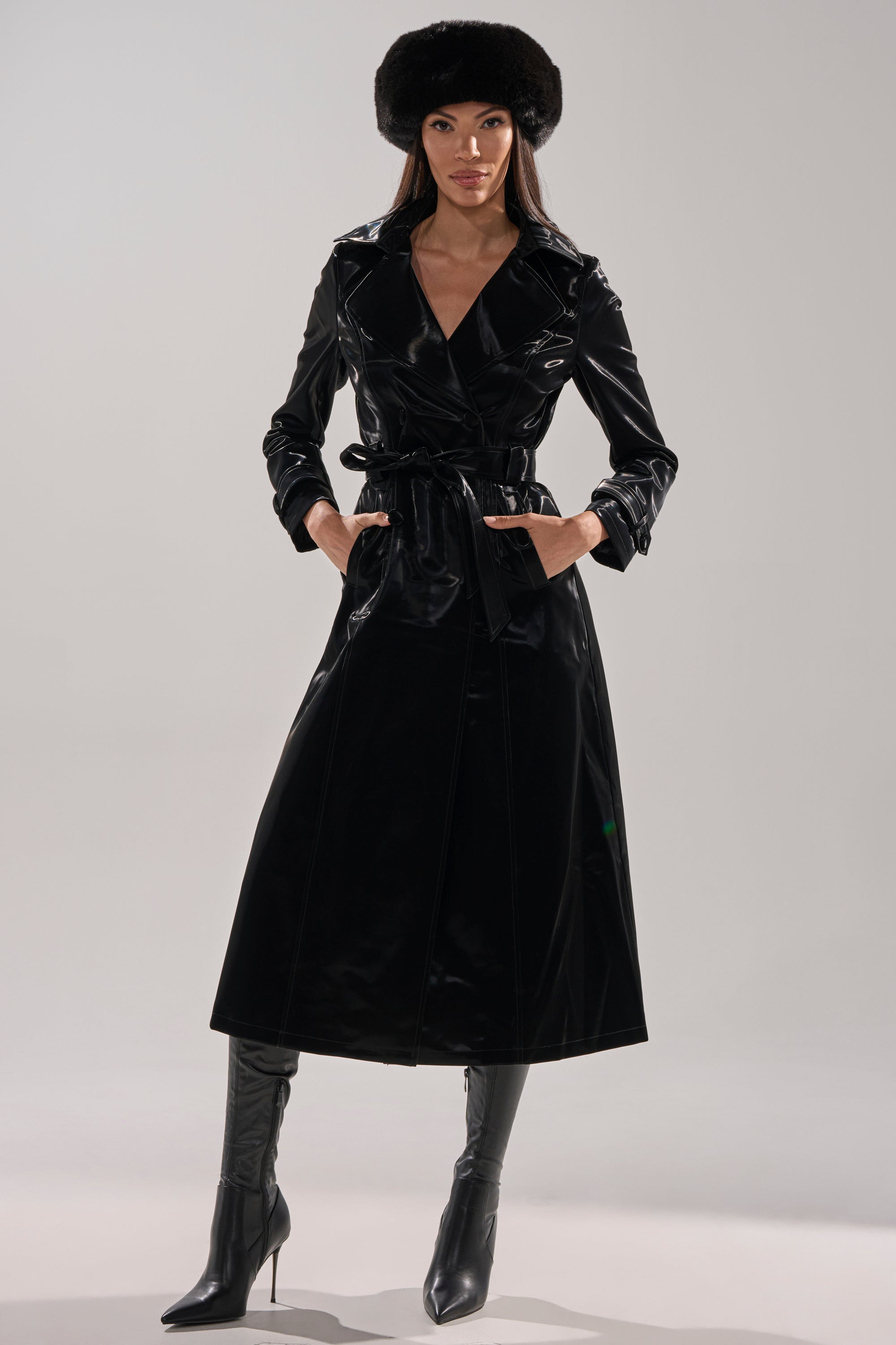 A woman wearing the SANDO LIQUID VELVET TRENCH IN BLACK, belted at the waist, stands with her hands in her pockets, paired with a black faux fur hat and knee-high black heeled boots against a light background.