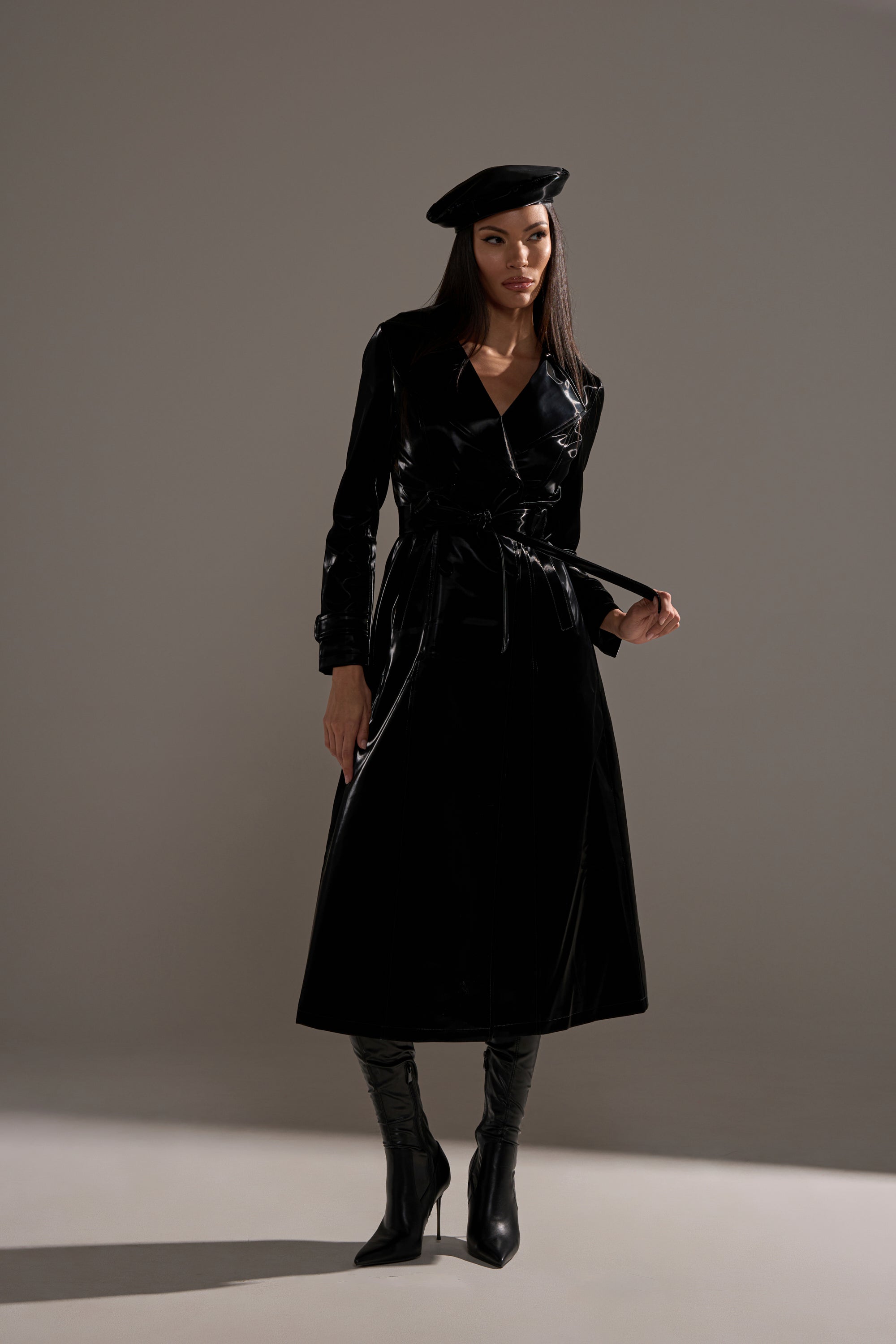 A woman stands against a plain gray background wearing a black beret, knee-high black heeled boots, and the SANDO LIQUID VELVET TRENCH IN BLACK with a longline silhouette.