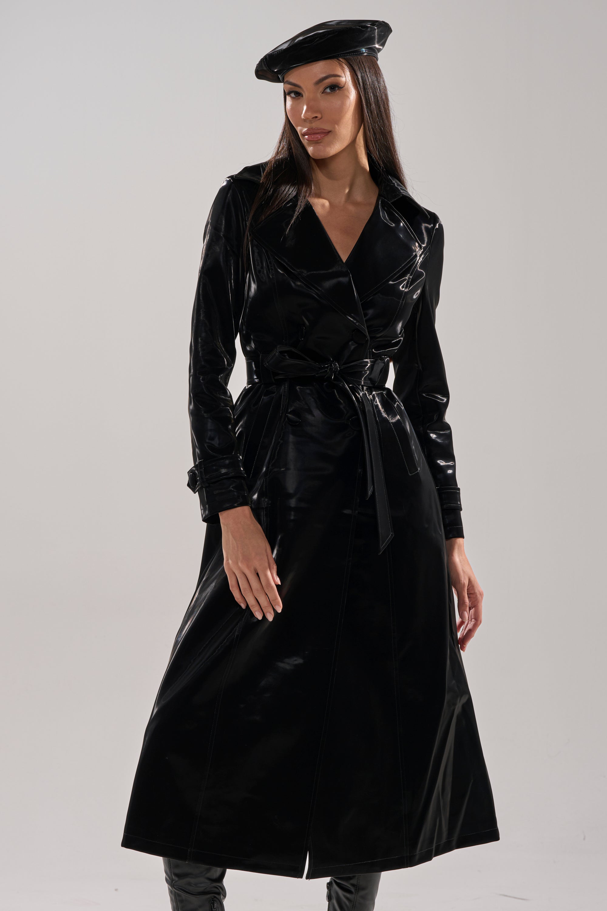 A woman stands confidently against a plain light gray background, wearing the SANDO LIQUID VELVET TRENCH IN BLACK, paired with a matching black beret and boots.