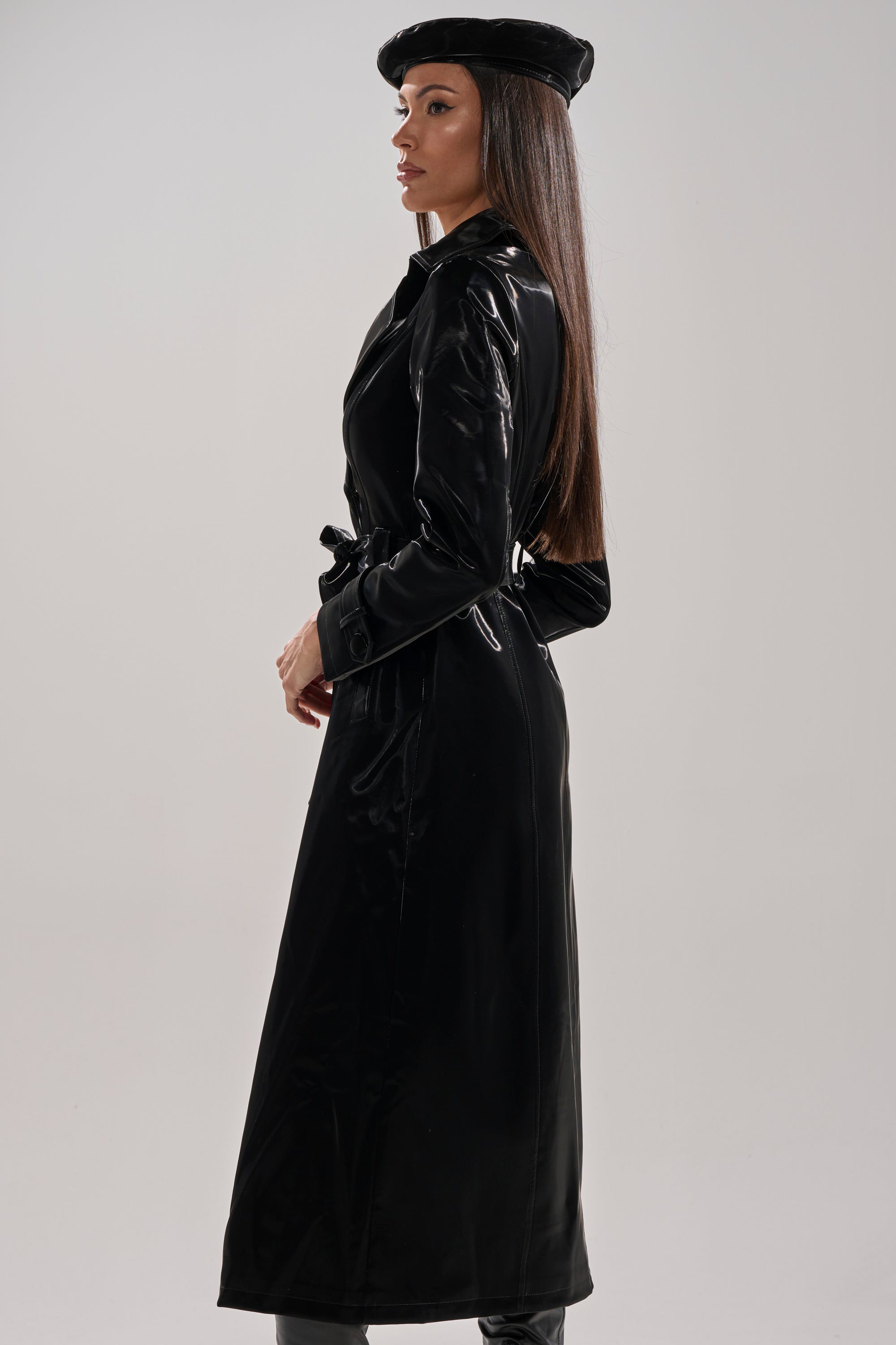 A woman with long straight hair stands in profile wearing the SANDO LIQUID VELVET TRENCH IN BLACK, a matching beret, and knee-high boots, set against a plain light background.