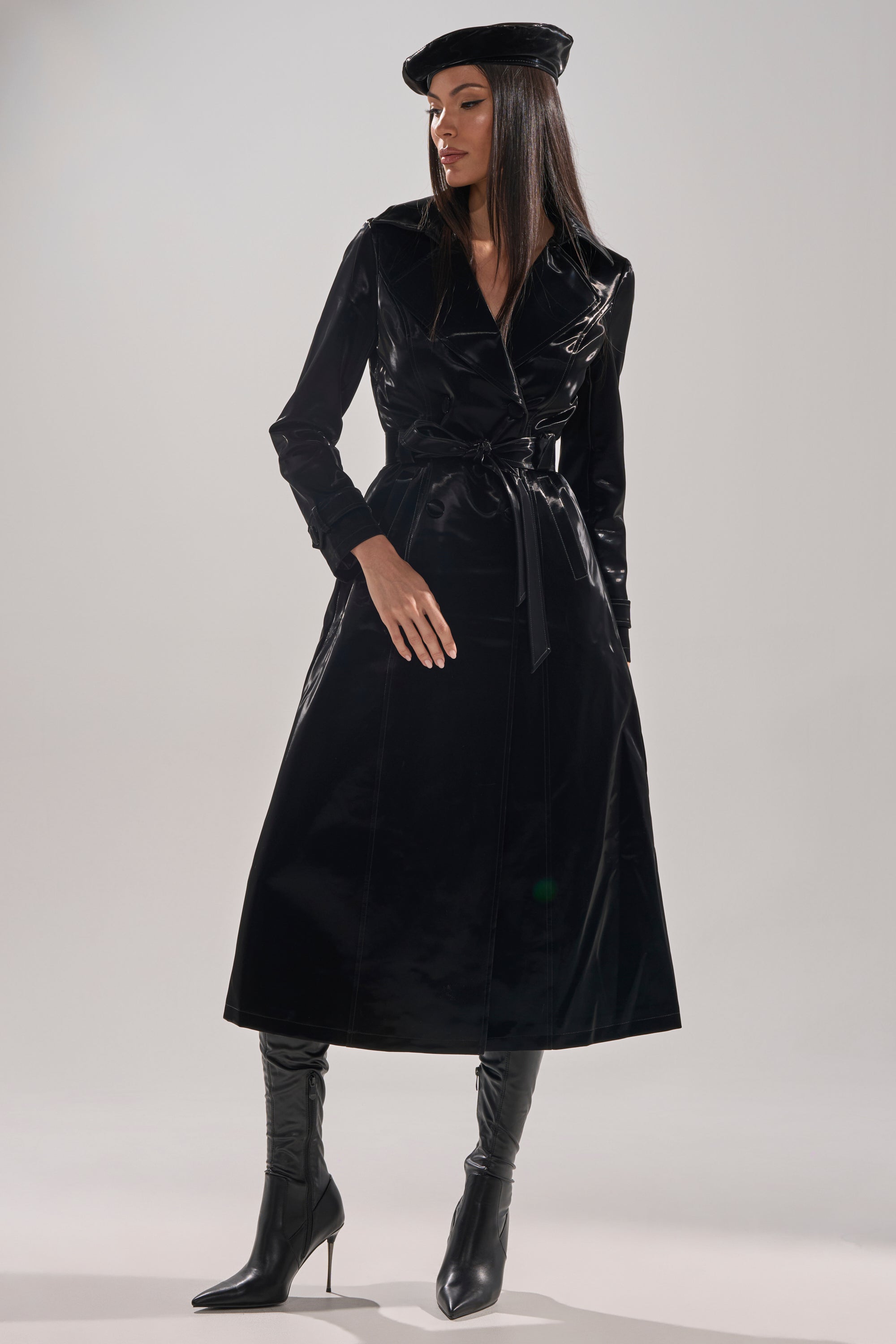 A woman models the SANDO LIQUID VELVET TRENCH IN BLACK, pairing it with knee-high boots and a black beret, as she poses confidently against a plain light background.