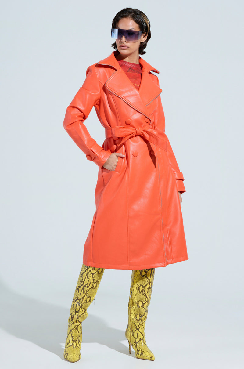 Clad in the SANDO ZIPPER TRIM TRENCH IN ORANGE from the AKIRA Label, a person exudes confidence as they pair it with yellow snake-print knee-high boots and large sunglasses, standing gracefully against a light background.