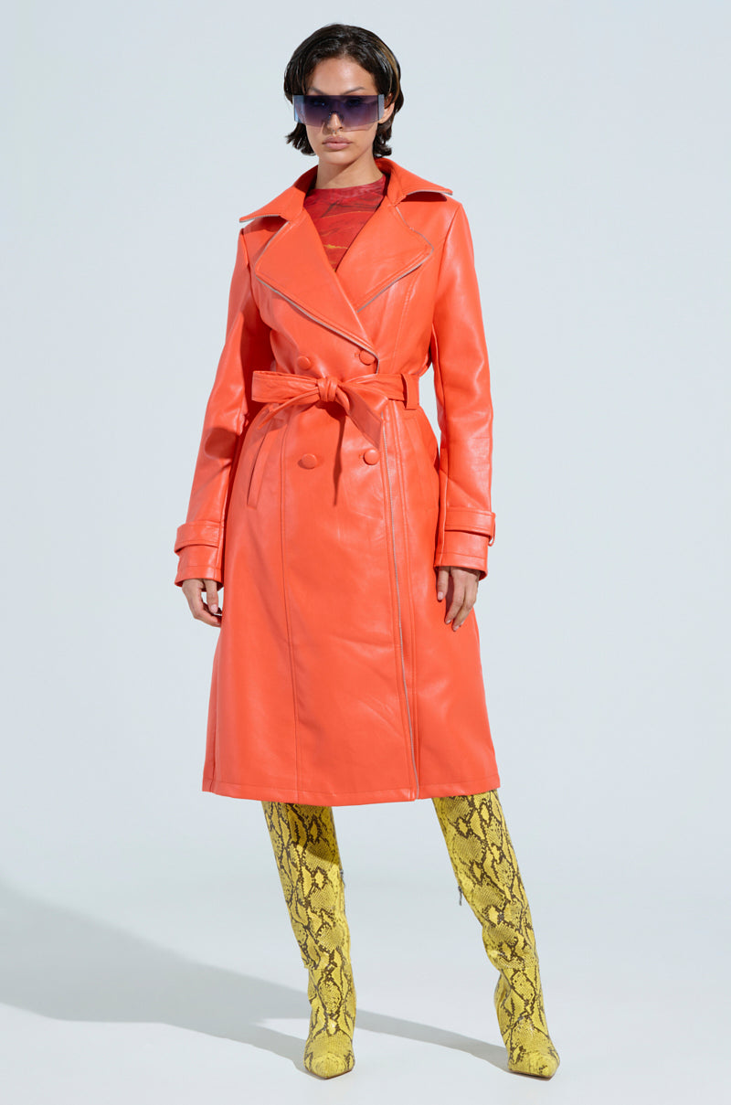 A person stands confidently wearing the SANDO ZIPPER TRIM TRENCH in orange, featuring a waist belt and zipper trim, paired with tall, textured yellow boots. They also wear large, dark sunglasses. The plain and light-colored background highlights the bold style reminiscent of the AKIRA Label aesthetic.