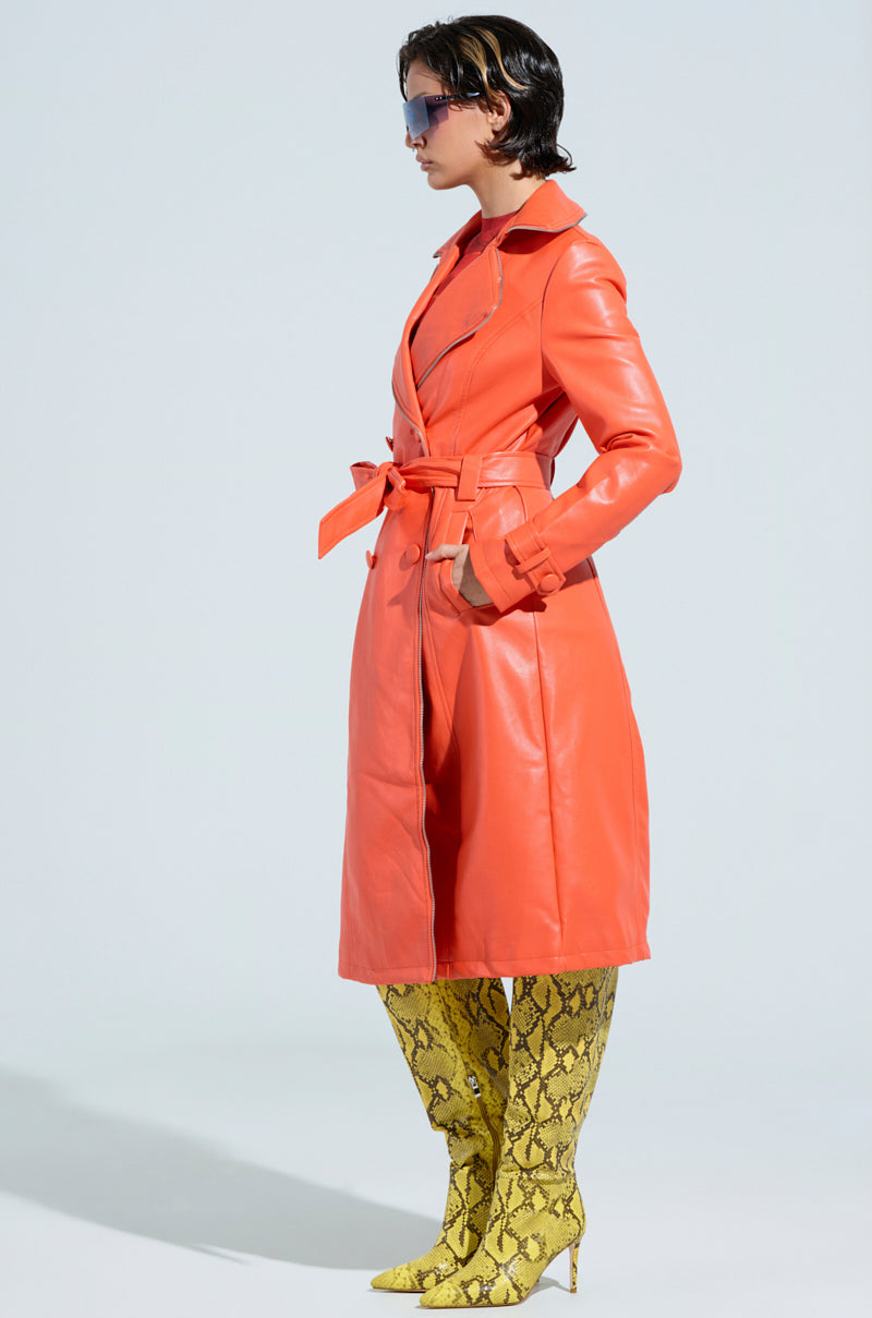 A person stands in profile against a light background, wearing the SANDO ZIPPER TRIM TRENCH IN ORANGE, tied at the waist. They are accessorized with large sunglasses and knee-high yellow snakeskin pattern boots, their short hair adding to the bold style reminiscent of the AKIRA Label.