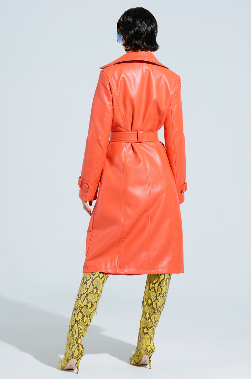 A person stands facing away, wearing the SANDO ZIPPER TRIM TRENCH IN ORANGE and yellow snake-print knee-high boots from the AKIRA Label. The background is light blue.