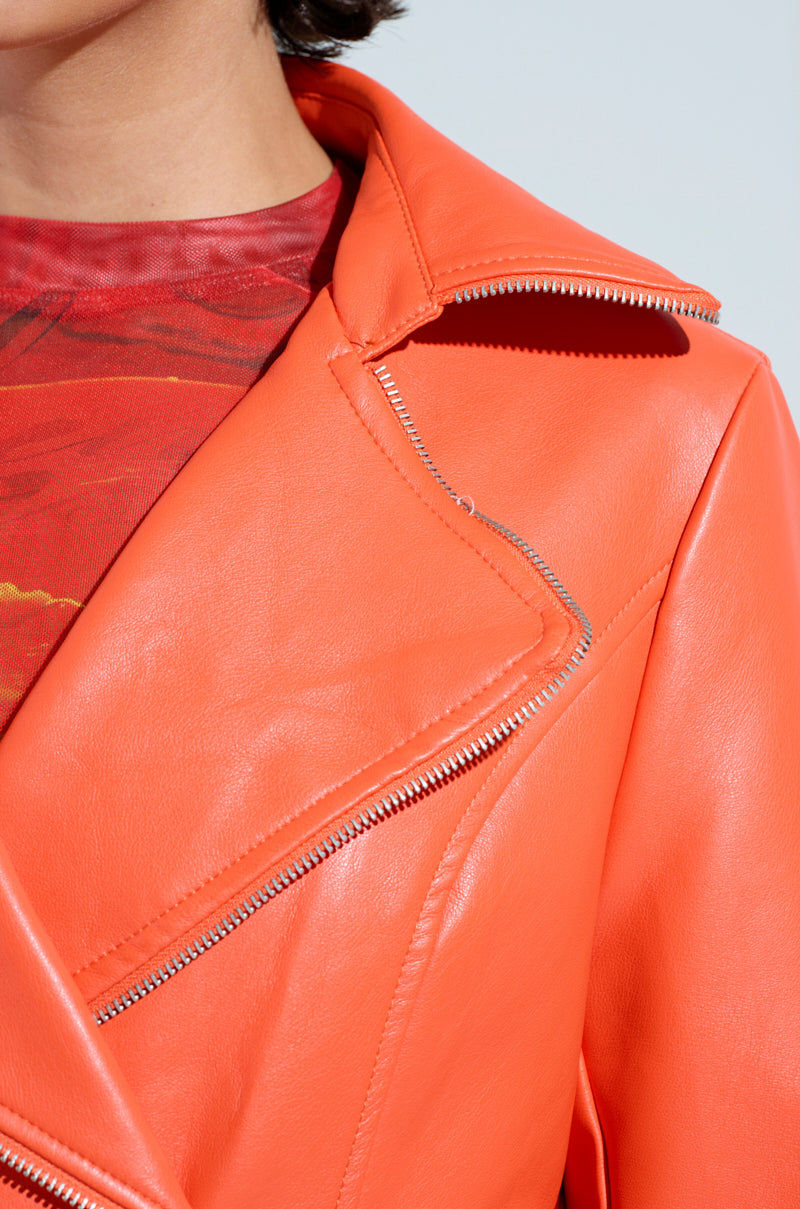 Close-up of a person wearing the SANDO ZIPPER TRIM TRENCH IN ORANGE by AKIRA Label, showcasing its silver zippers over a red patterned top. The trench coat features a notched lapel, focusing on the shoulder and chest area.