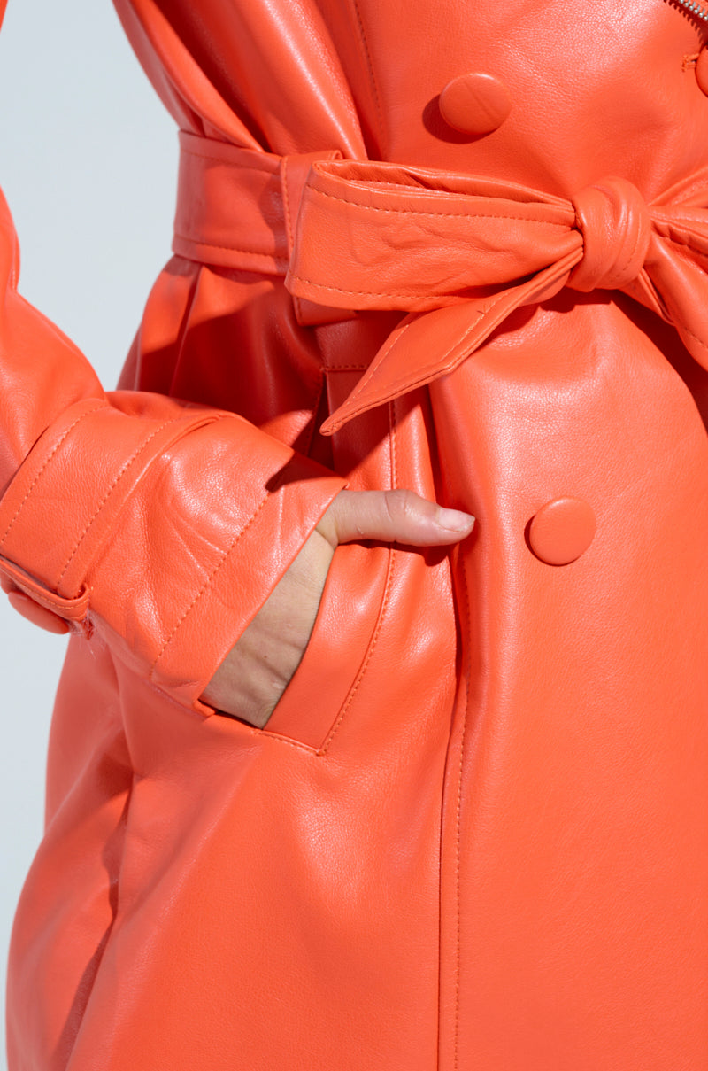 A person is wearing the SANDO ZIPPER TRIM TRENCH IN ORANGE, featuring a tied belt and large buttons. One hand is tucked into a side pocket. The glossy finish of the faux leather is highlighted in the bright lighting, enhancing its chic appearance.