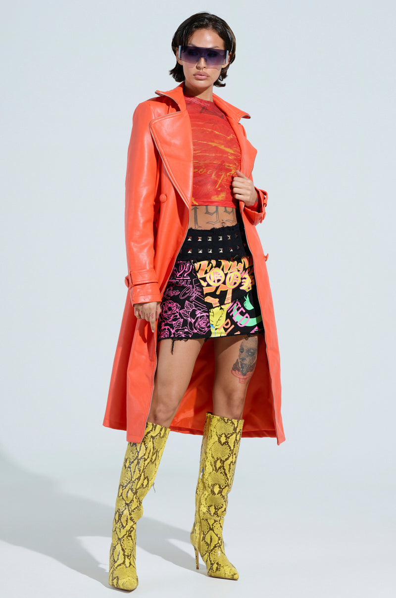 A person confidently stands against a plain background, dressed in the AKIRA Label SANDO Zipper Trim Trench in Orange, paired with a red patterned shirt, black shorts featuring colorful designs, yellow snake-patterned knee-high boots, and sunglasses.