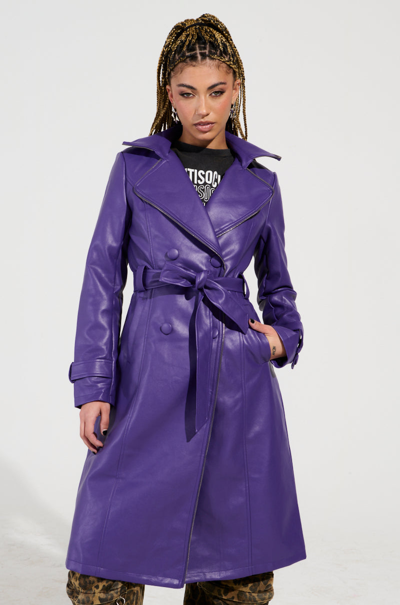 An individual with braided hair confidently stands against a plain background, wearing the vibrant SANDO Zipper Trim Trench in Purple. The faux leather trench coat features a zipper trim and a waist-cinching belt, elegantly revealing a black graphic T-shirt and patterned pants underneath.