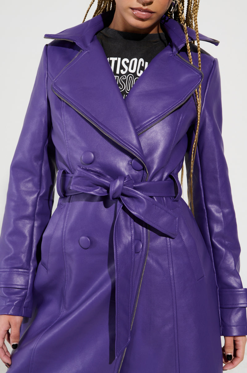 A person wearing the SANDO Zipper Trim Trench in purple, a stylish faux leather coat with a waist-cinching tied belt and substantial lapels. The coat is adorned with double-breasted buttons, and the person has long, braided hair. The sunlight accentuates the texture of the faux leather.
