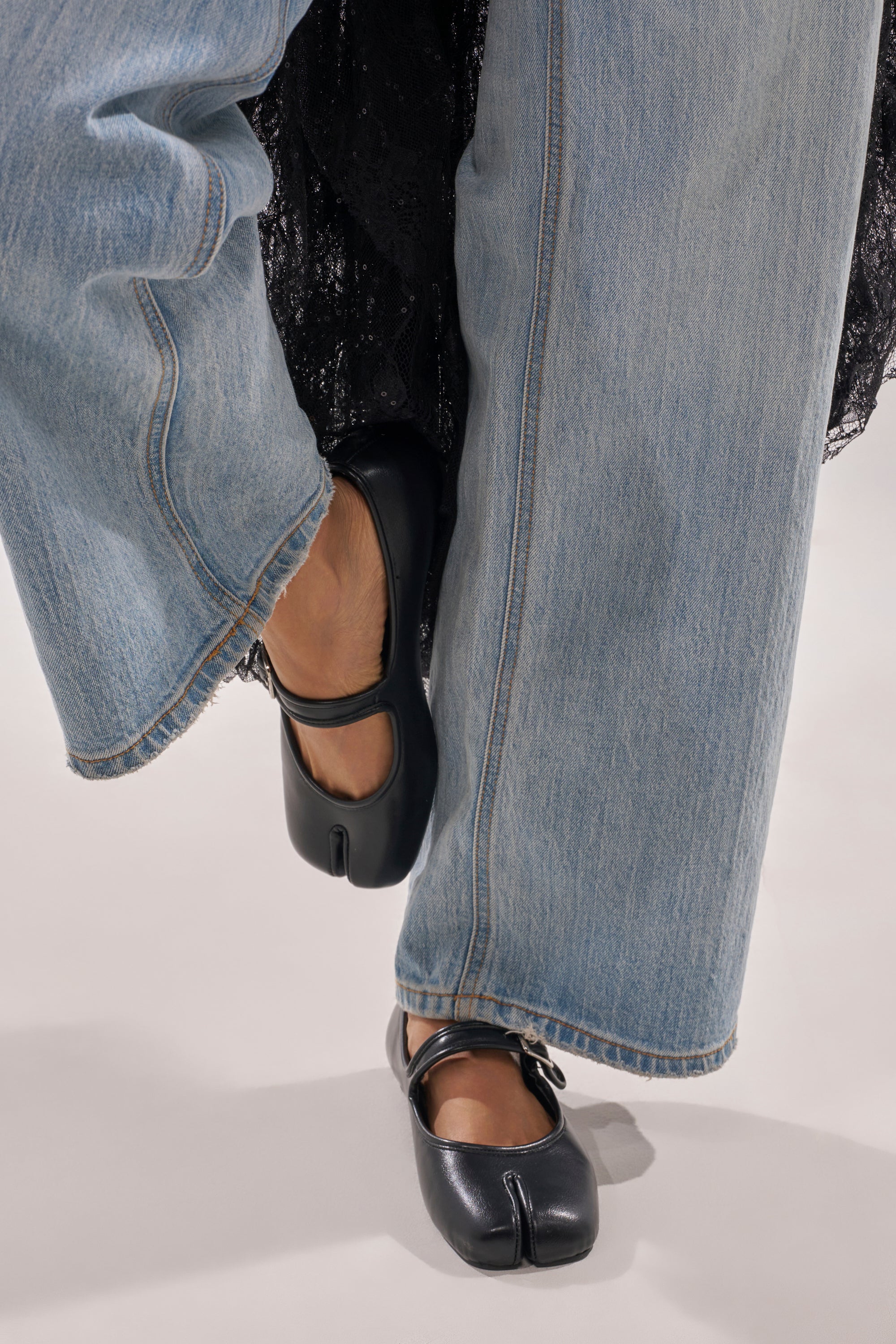 Wearing light blue wide-leg jeans, a person stands with one foot slightly ahead, showcasing the AKIRA GOLD SANG BLACK PU SPLIT TOE FLAT with comfort cushion insoles. 0