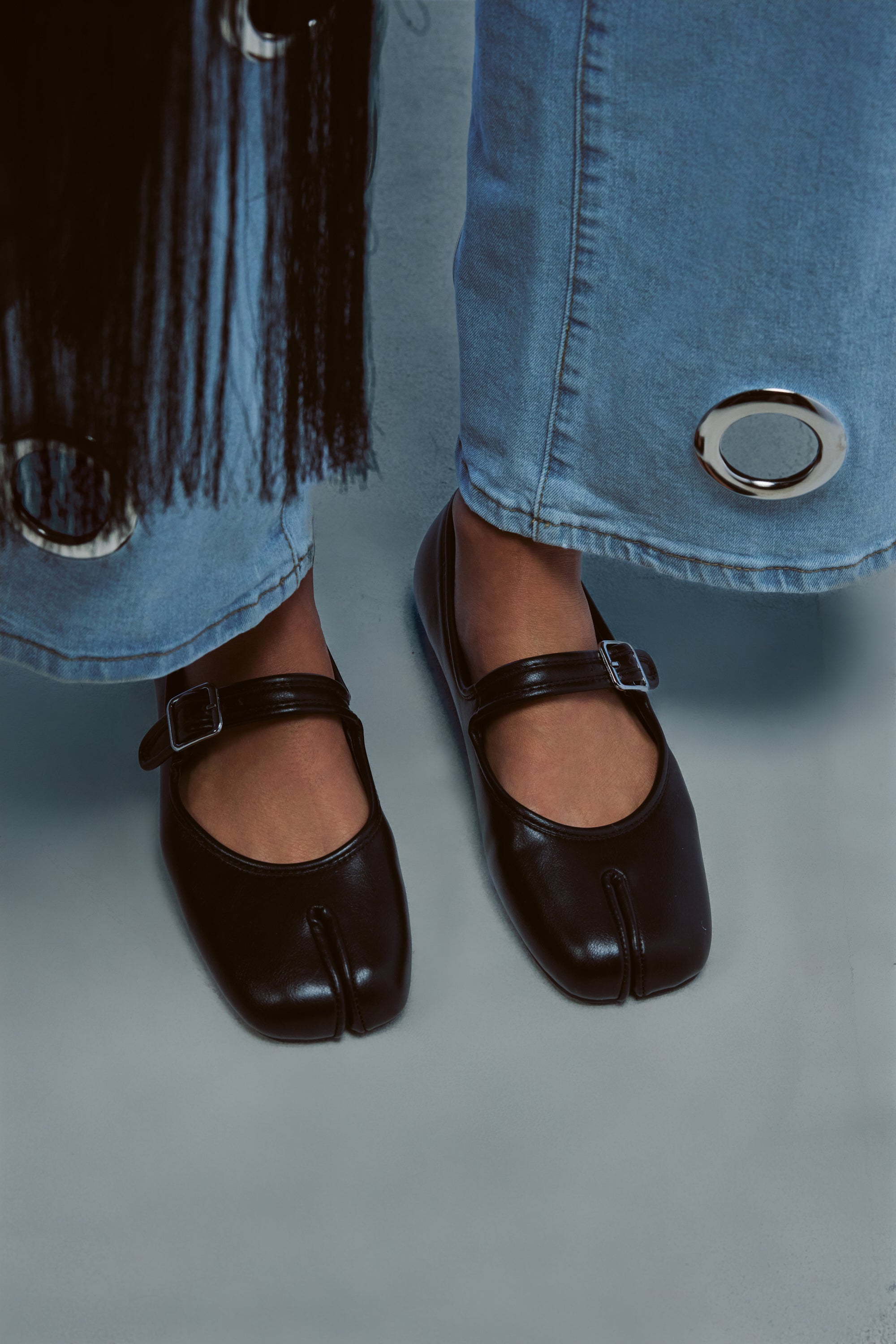A person wearing the AKIRA GOLD SANG BLACK PU SPLIT TOE FLAT and light blue jeans with large metal grommets stands on a gray floor.