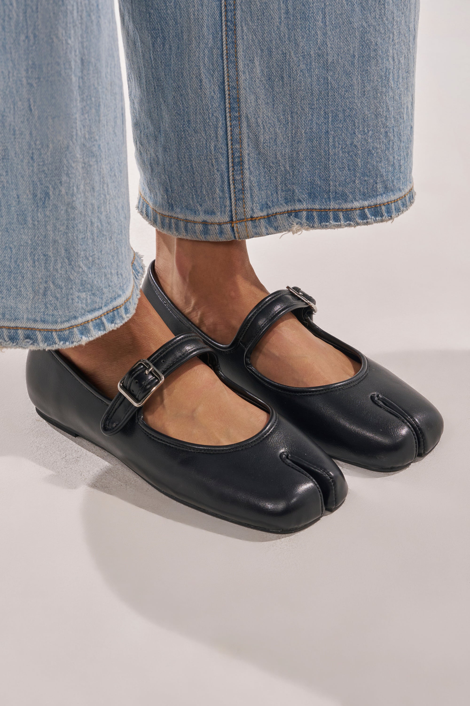 Wearing cropped light blue jeans, a person stands on a light surface in AKIRA GOLD SANG BLACK PU SPLIT TOE FLAT shoes featuring a faux leather finish and distinctive split square toe design. 1