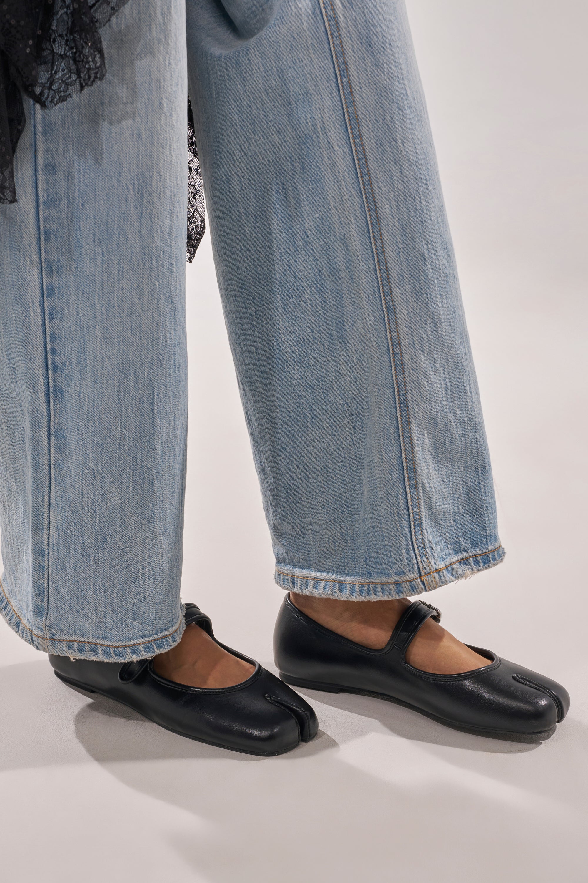 Someone wearing light blue wide-leg jeans and the AKIRA GOLD SANG BLACK PU SPLIT TOE FLAT—faux leather Mary Jane shoes with a split square toe—stands on a light-colored surface. 3
