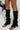 A person wearing black knit leg warmers over the AZALEA WANG SAPPHIRA BLACK SANDAL with stiletto heels, holding a black handbag. The focus is on the legs and footwear against a plain background.