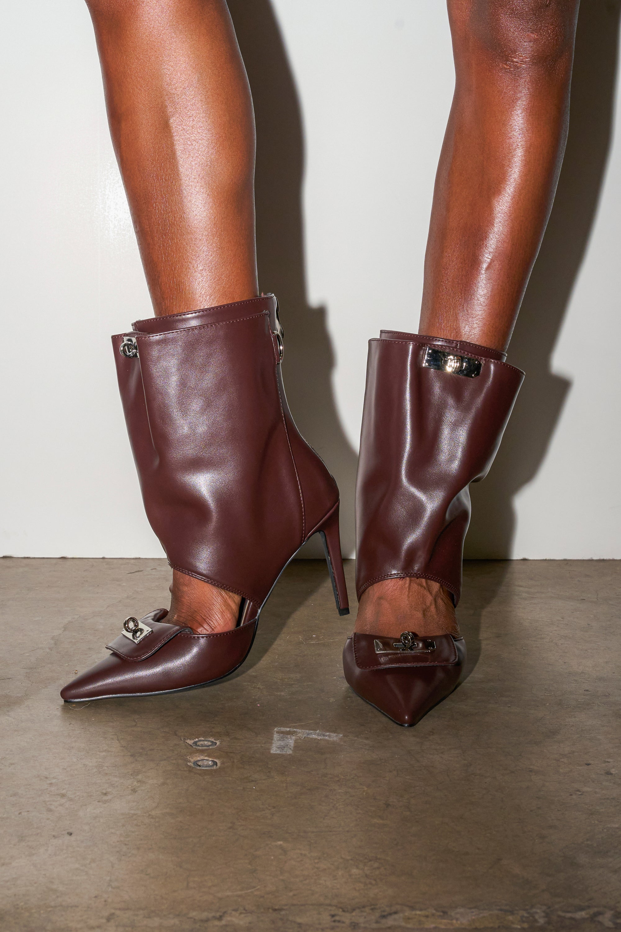 A person wears the AZALEA WANG SARVESH BROWN CUTOUT STILETO BOOTIE, featuring shiny dark brown pointed toes, boot-like leather coverings, and metallic buckles, while standing on a concrete floor against a white wall. 0
