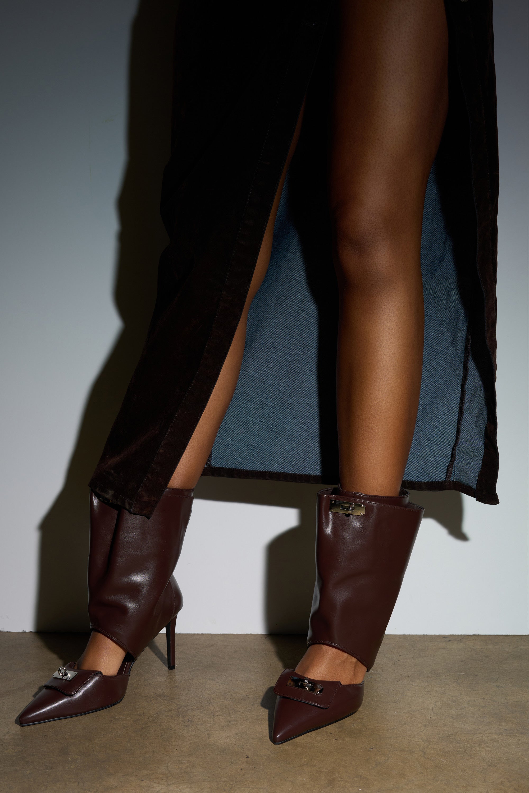 Wearing a dark skirt with a high slit, someone showcases bare legs and the AZALEA WANG SARVESH BROWN CUTOUT STILETTO BOOTIE while standing on a concrete floor against a plain background.