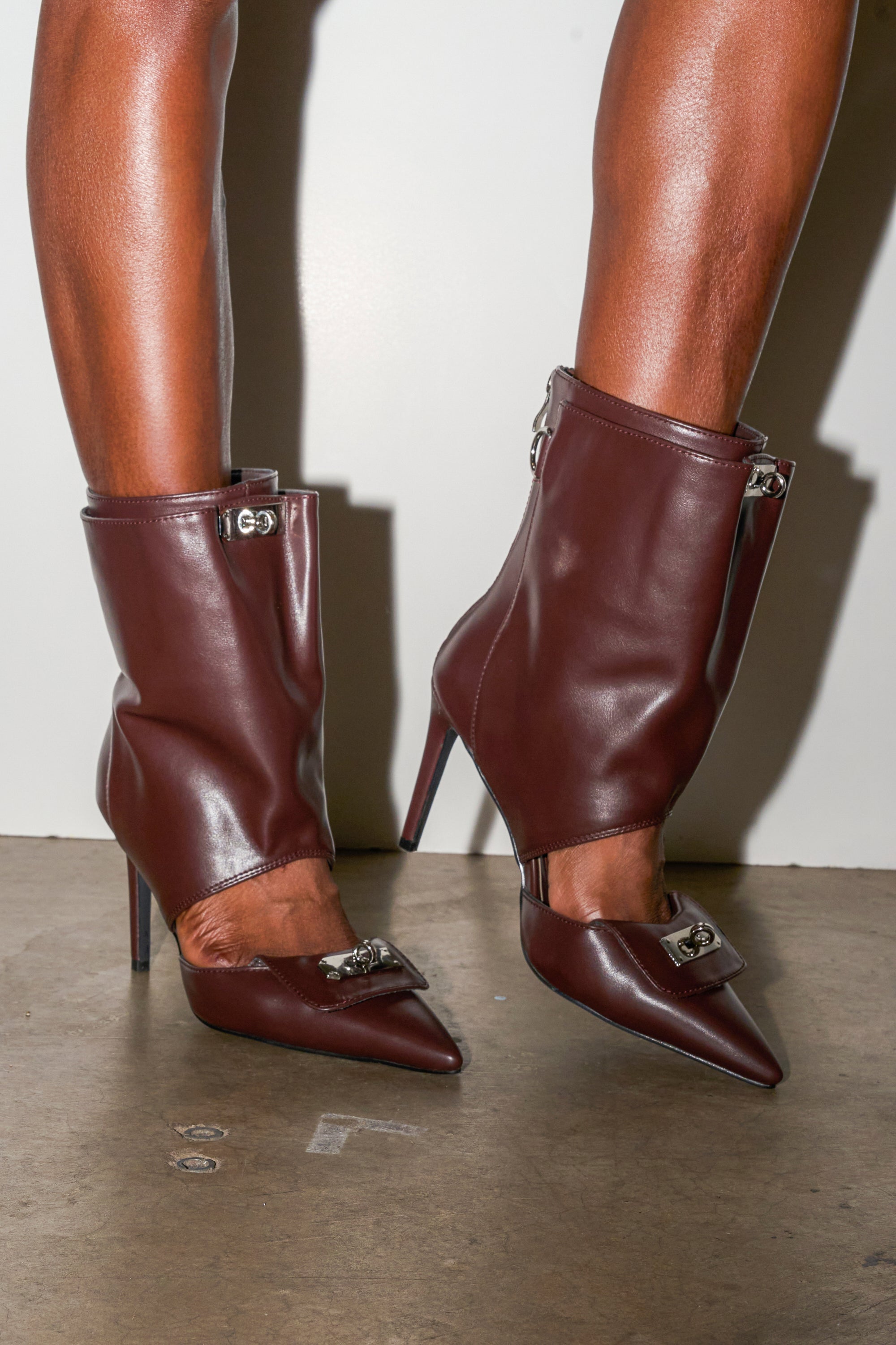 The AZALEA WANG SARVESH BROWN CUTOUT STILETTO BOOTIE is shown on brown-skinned legs against a plain background, featuring pointed toes, stylish cutouts, and sleek stiletto heels. 1