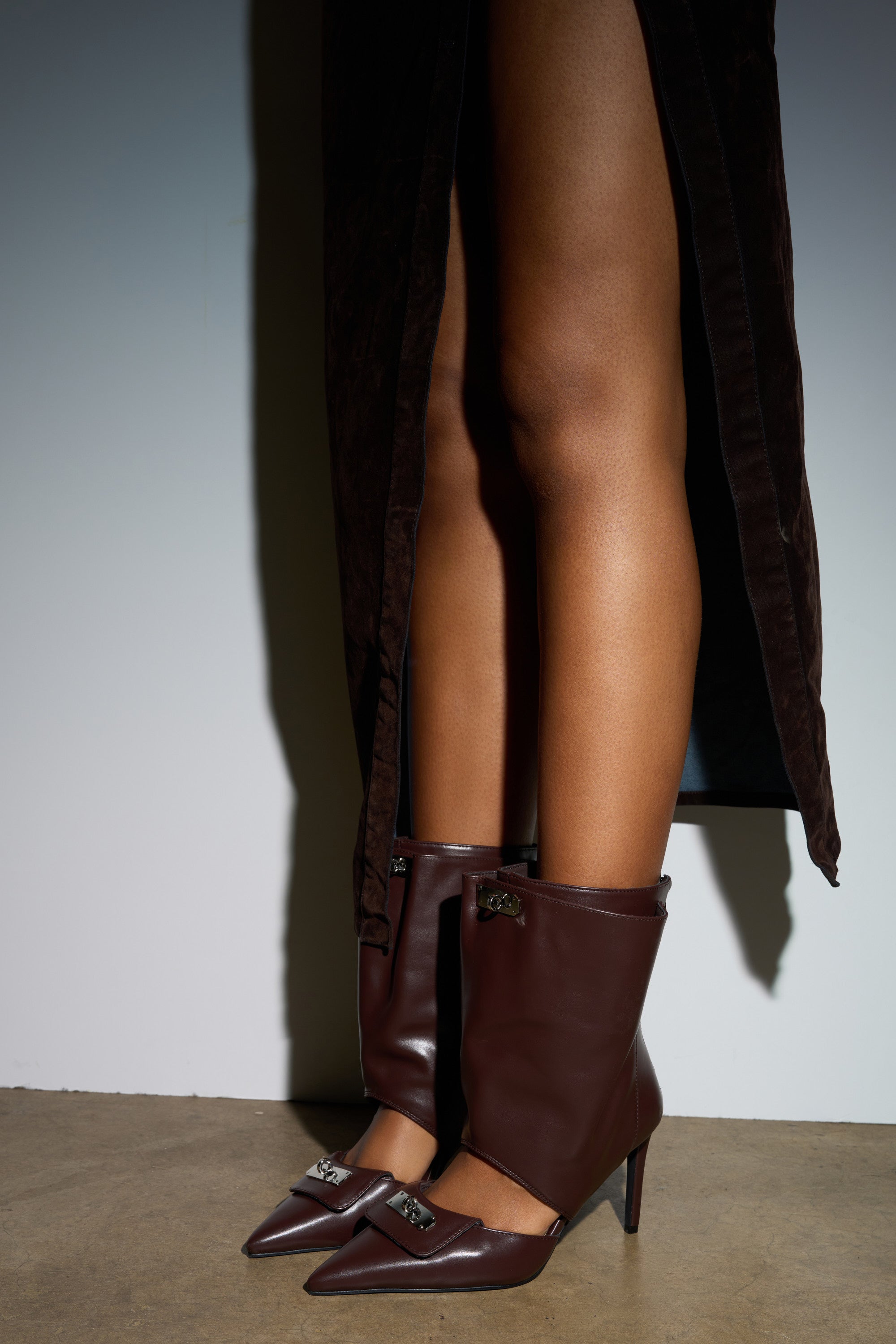 Indoors, a person wears the AZALEA WANG SARVESH BROWN CUTOUT STILETTO BOOTIE with a dark brown skirt featuring a front slit, showcasing bare legs and the bootie's metallic embellishments.