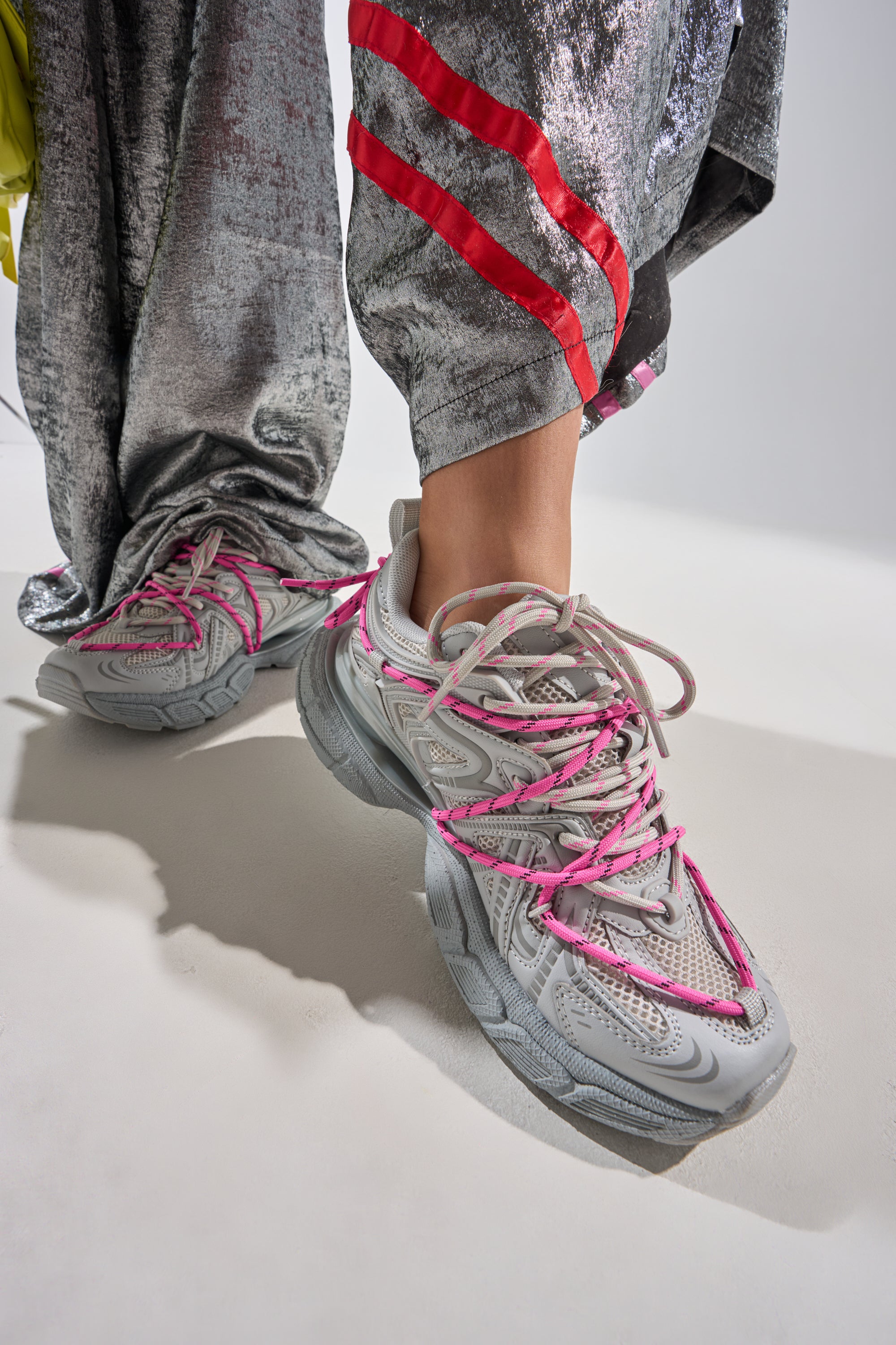 A person in shiny silver pants with red stripes stands wearing AZALEA WANG SATINE GREY LACE UP SNEAKERS, which feature a chunky sculpted sole and pink laces, on a white background. 0