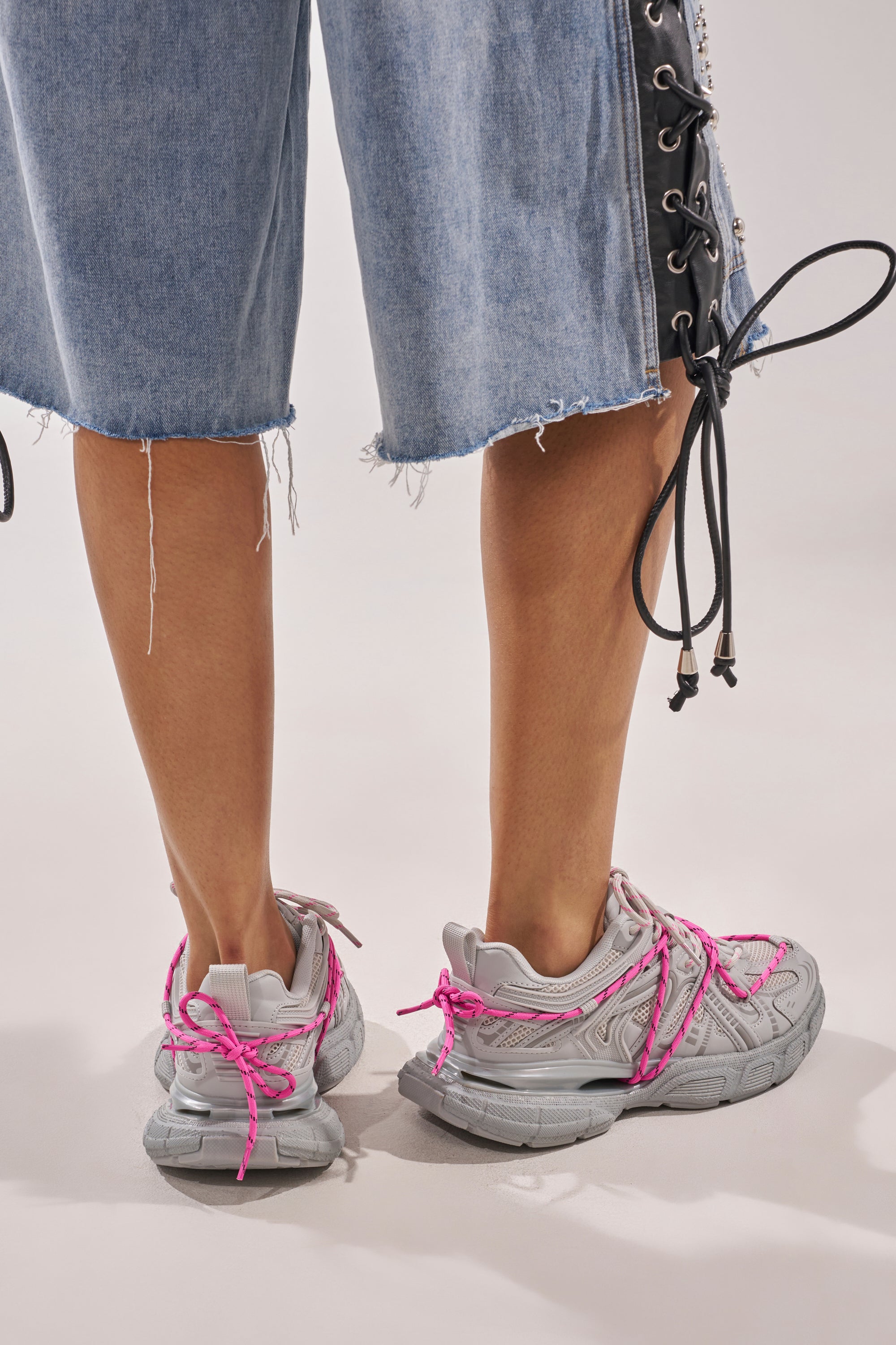 A person shown from the back wears frayed denim shorts with lace-up sides and AZALEA WANG SATINE GREY LACE UP SNEAKER, featuring bright pink laces, against a neutral background. 3