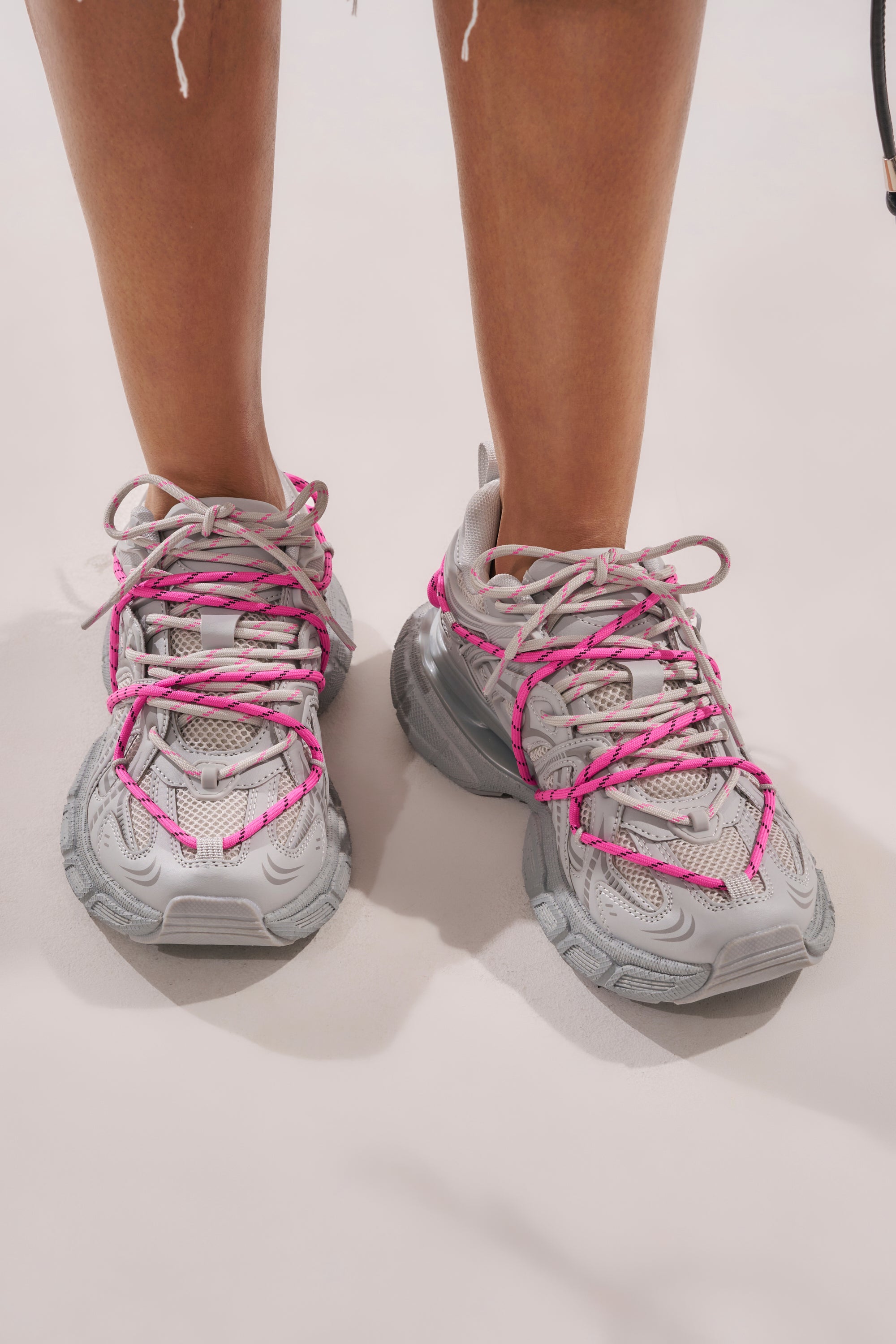 A person stands on a light surface, visible from mid-calf down, wearing AZALEA WANG SATINE GREY LACE UP SNEAKERS with chunky soles and bright pink laces. 4