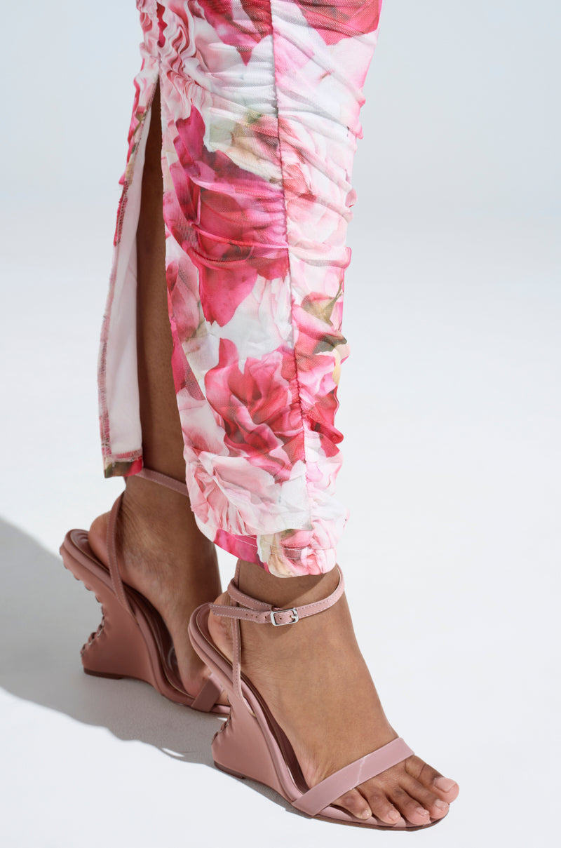 Close-up of a person wearing floral-patterned pants and the AZALEA WANG SAWNEY MAUVE LACE UP PATENT SANDALS with jagged soles. The pants have ruched details and a side slit, adding elegance against the light gray background.