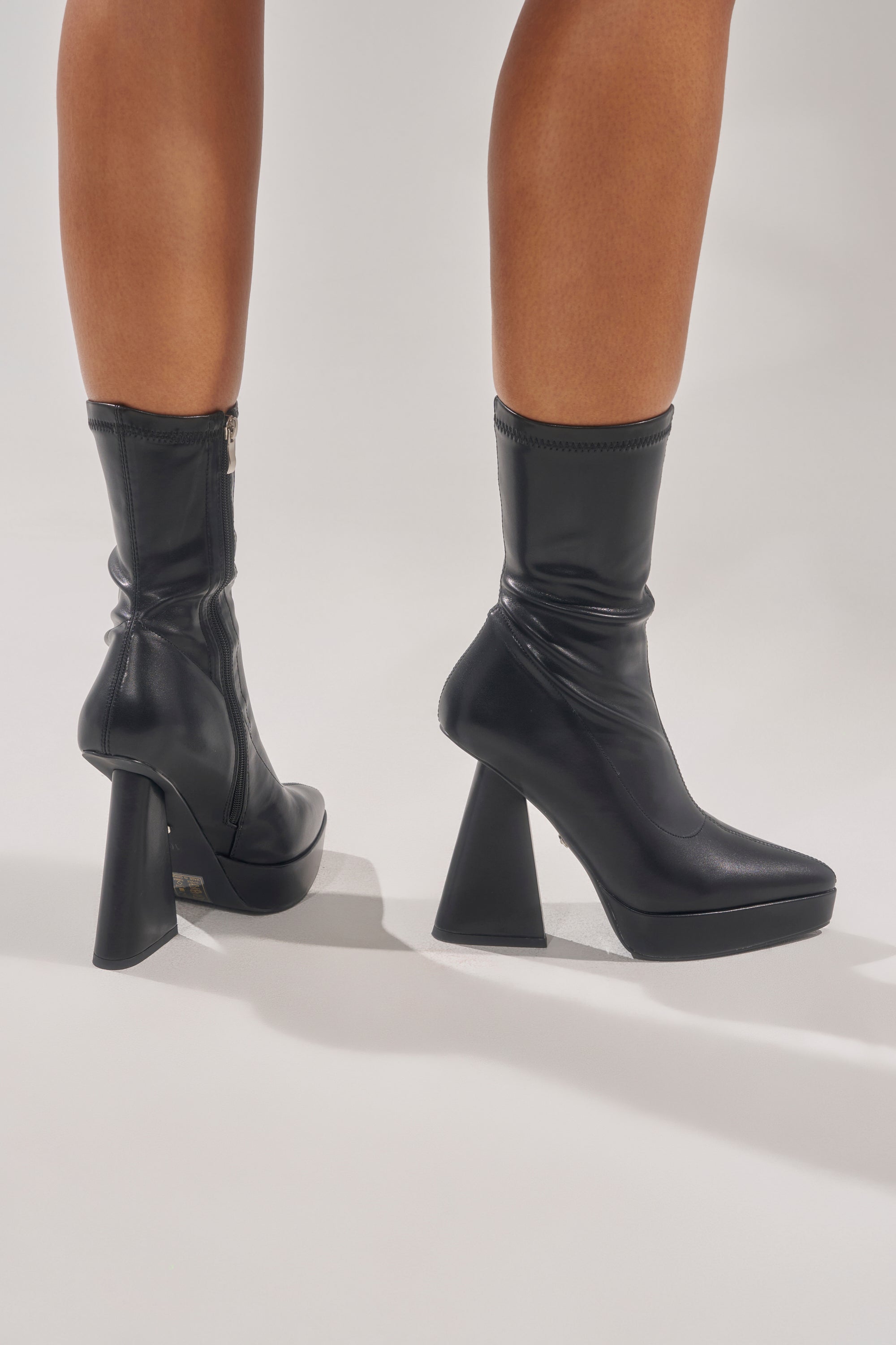 A person stands on a light background wearing the AZALEA WANG SAXTON BLACK PU CHUNKY POINTED TOE BOOTIE, featuring geometric chunky heels, side zippers, and a sleek, modern design. 0