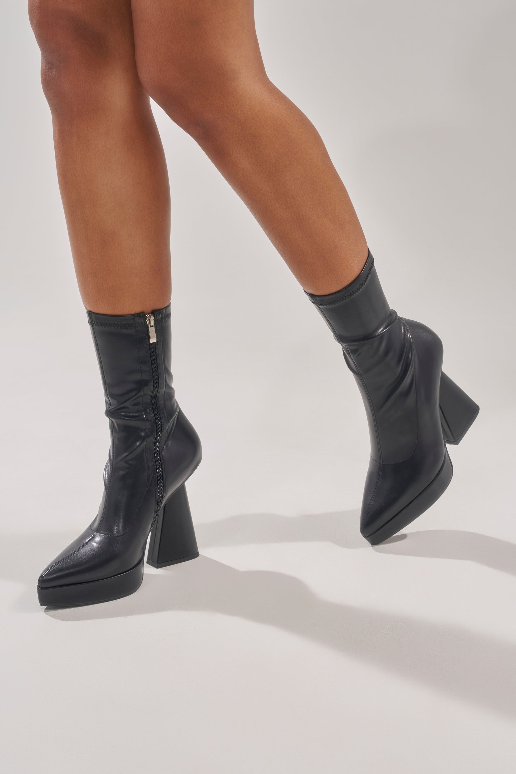 A person wearing the AZALEA WANG SAXTON BLACK PU CHUNKY POINTED TOE BOOTIE with side zippers and platform soles stands on a white background, one leg slightly raised. 3