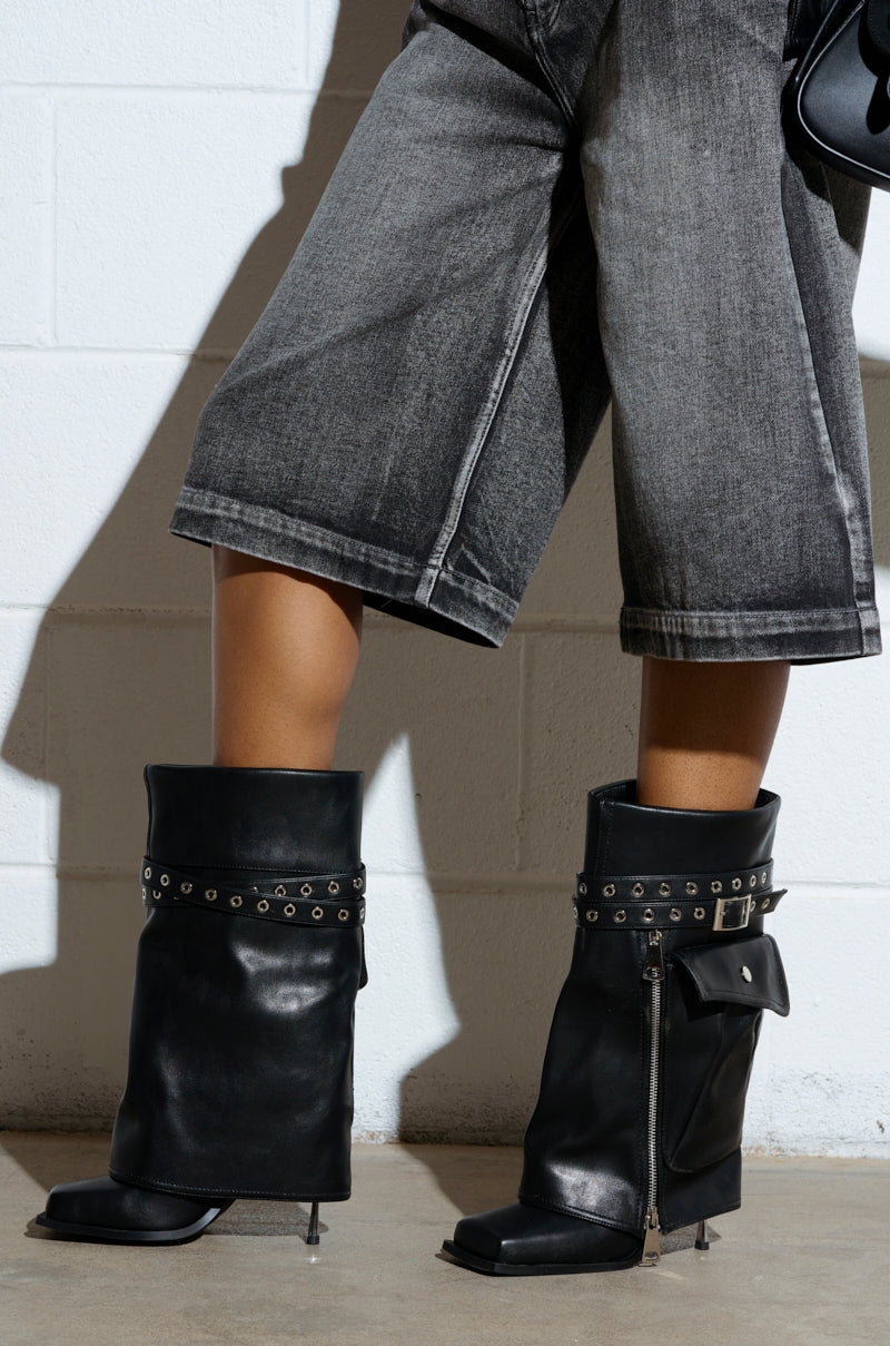 Sporting the AZALEA WANG SAYLAH BLACK FOLD OVER STILETTO BOOTIE with studded straps and zipper details, paired with wide-leg faded black jeans, a person stands perfectly complemented by the light-colored brick wall backdrop.