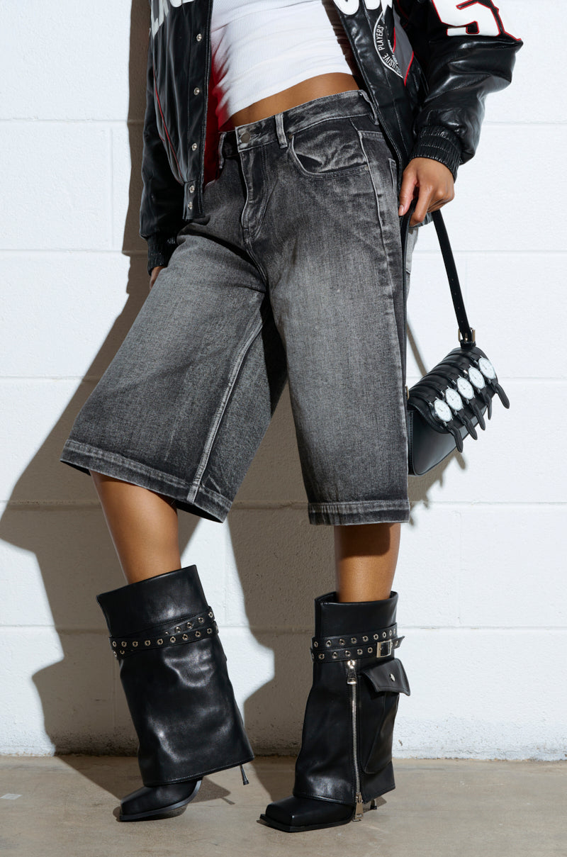 A person wearing distressed gray denim shorts, a partially visible black and red jacket, and AZALEA WANG SAYLAH BLACK FOLD OVER STILETTO BOOTIE poses against a white wall with a small black handbag hanging from their shoulder.