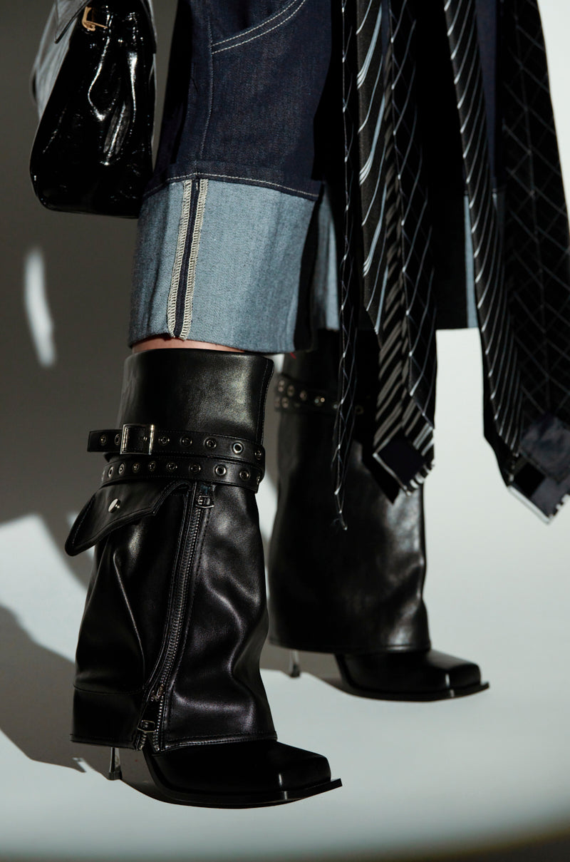 In a close-up, someone wears AZALEA WANG SAYLAH BLACK FOLD OVER STILETTO BOOTIE, their faux leather finish gleaming, paired with rolled-up jeans and a striped tie-detail top. A black bag partially appears on the left.