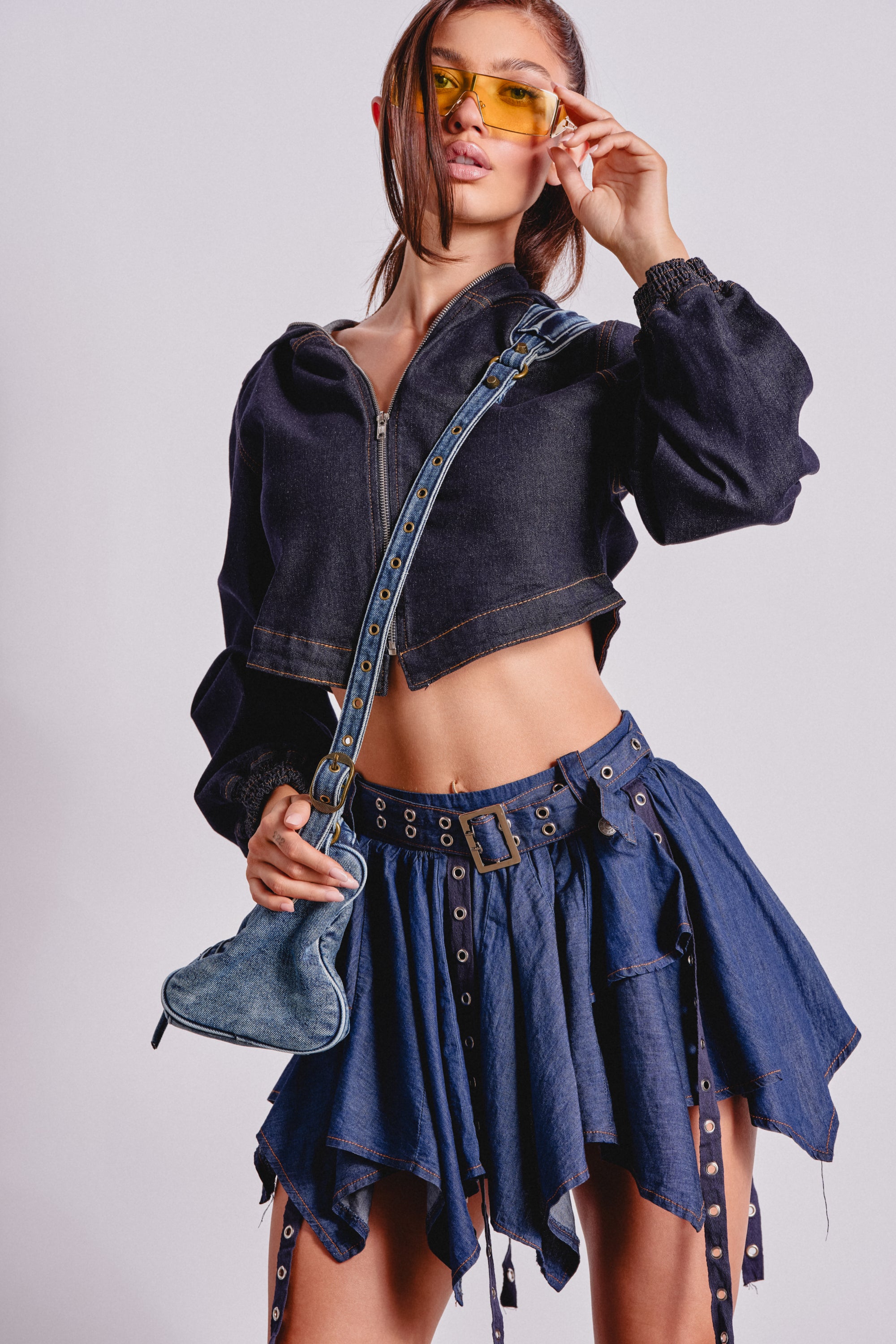 A woman poses in the BEAM ME UP HOODED DENIM JACKET, a blue asymmetrical denim skirt with grommet details, yellow-tinted sunglasses, and a blue guitar-shaped crossbody bag. 0