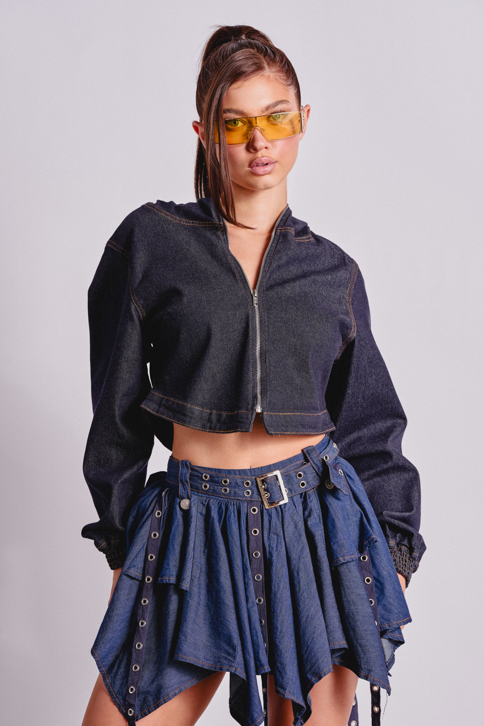 A young woman poses confidently in the BEAM ME UP HOODED DENIM JACKET, a high-waisted asymmetric denim skirt with grommet details, and yellow-tinted sunglasses, showcasing an effortless streetwear style against a plain light background. 2