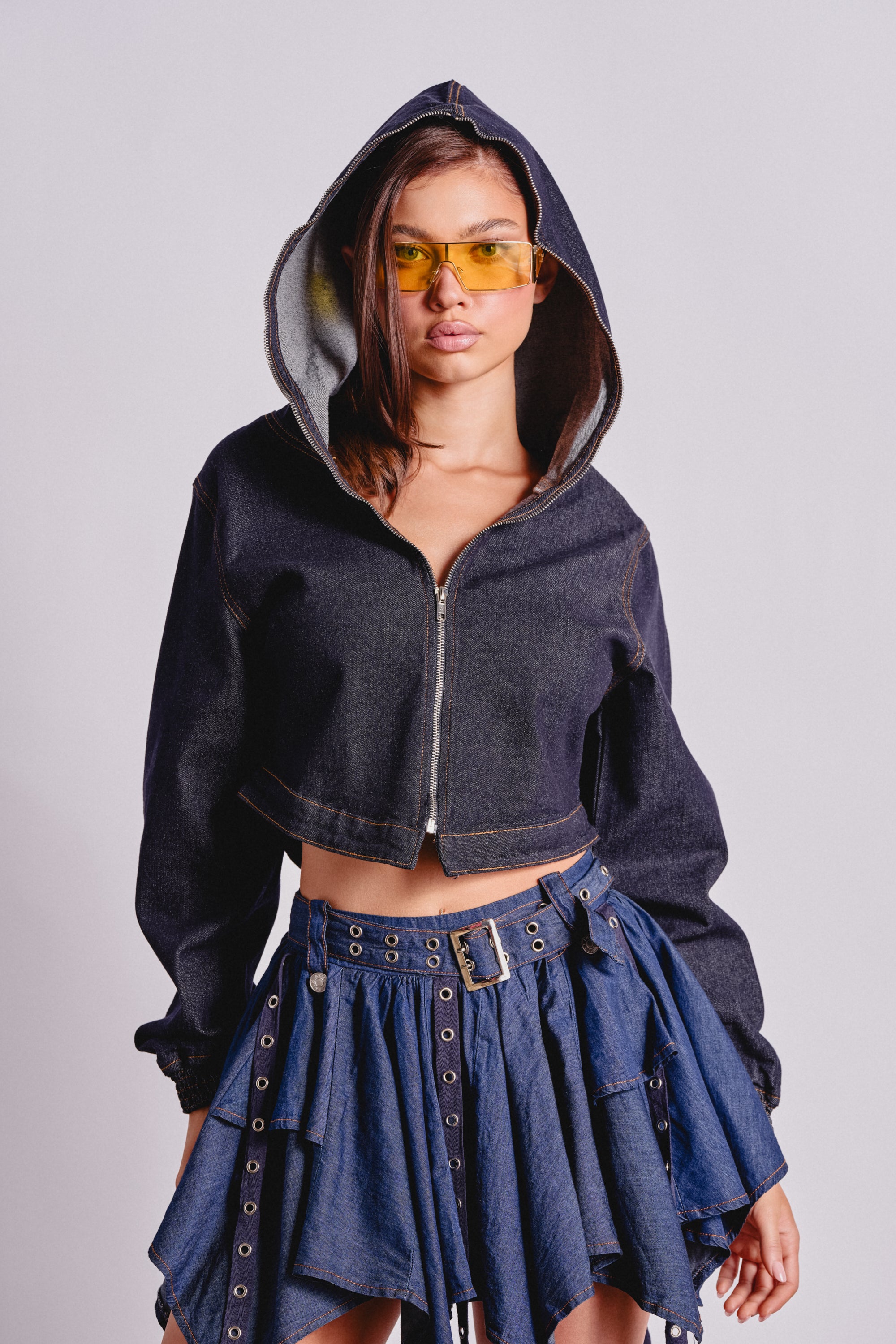 A woman stands facing the camera in the BEAM ME UP HOODED DENIM JACKET with the hood up, yellow-tinted sunglasses, and a pleated denim skirt, channeling effortless streetwear style against a plain light background. 3