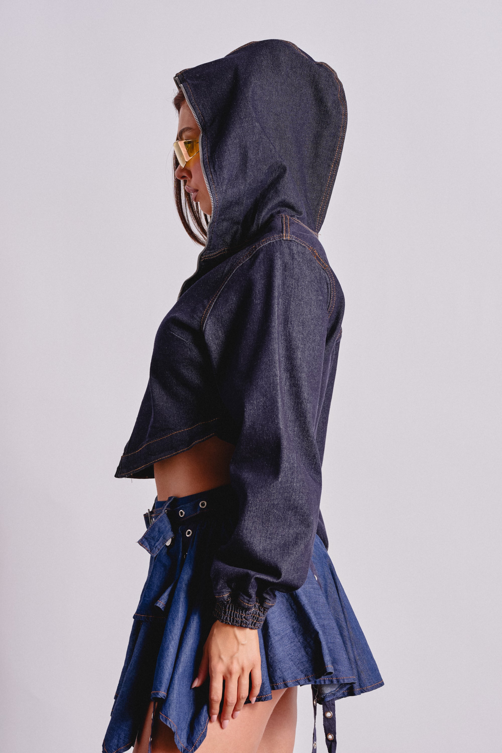 A woman stands in profile against a plain light background, wearing the BEAM ME UP HOODED DENIM JACKET with the hood up, yellow sunglasses, and a blue denim mini skirt with belt details. 4