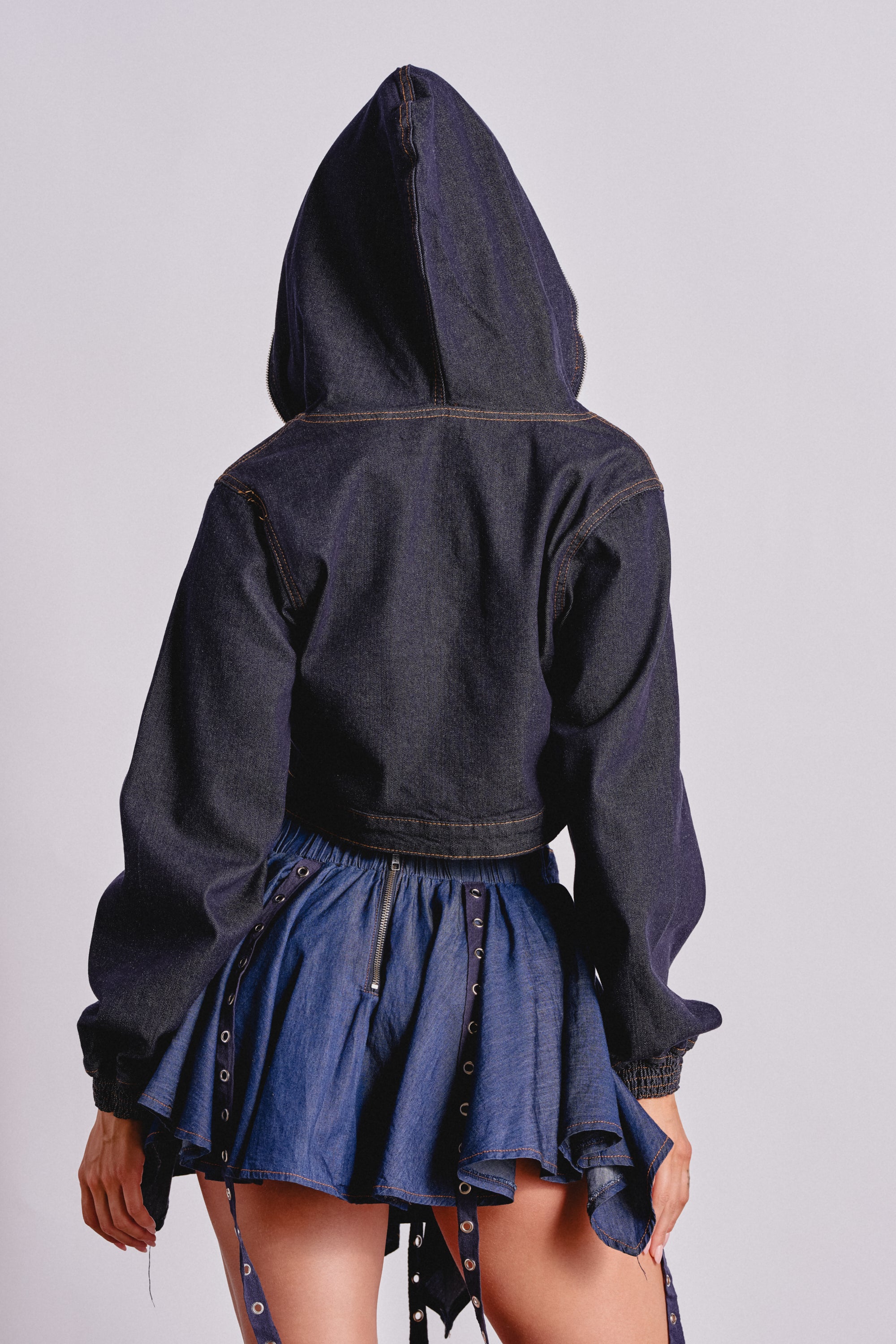 A person stands facing away, wearing the BEAM ME UP HOODED DENIM JACKET and a short, pleated blue denim skirt with grommet and strap details, showcasing bold streetwear style against a plain light background. 5
