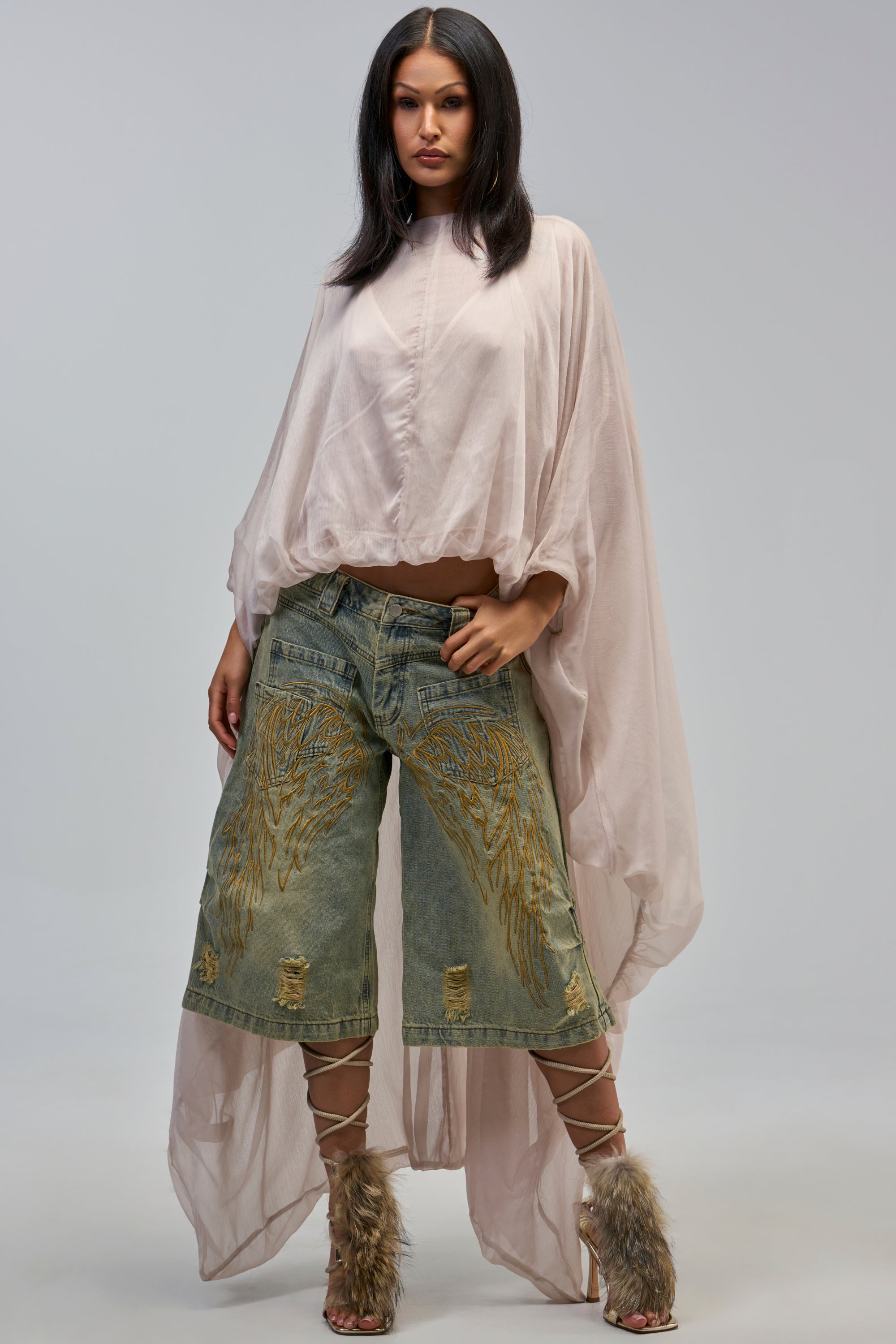 A woman poses confidently in a flowing sheer pale pink cape top, QUITE ANGELIC WASHED DENIM BERMUDA SHORTS, and lace-up heels with fur accents.