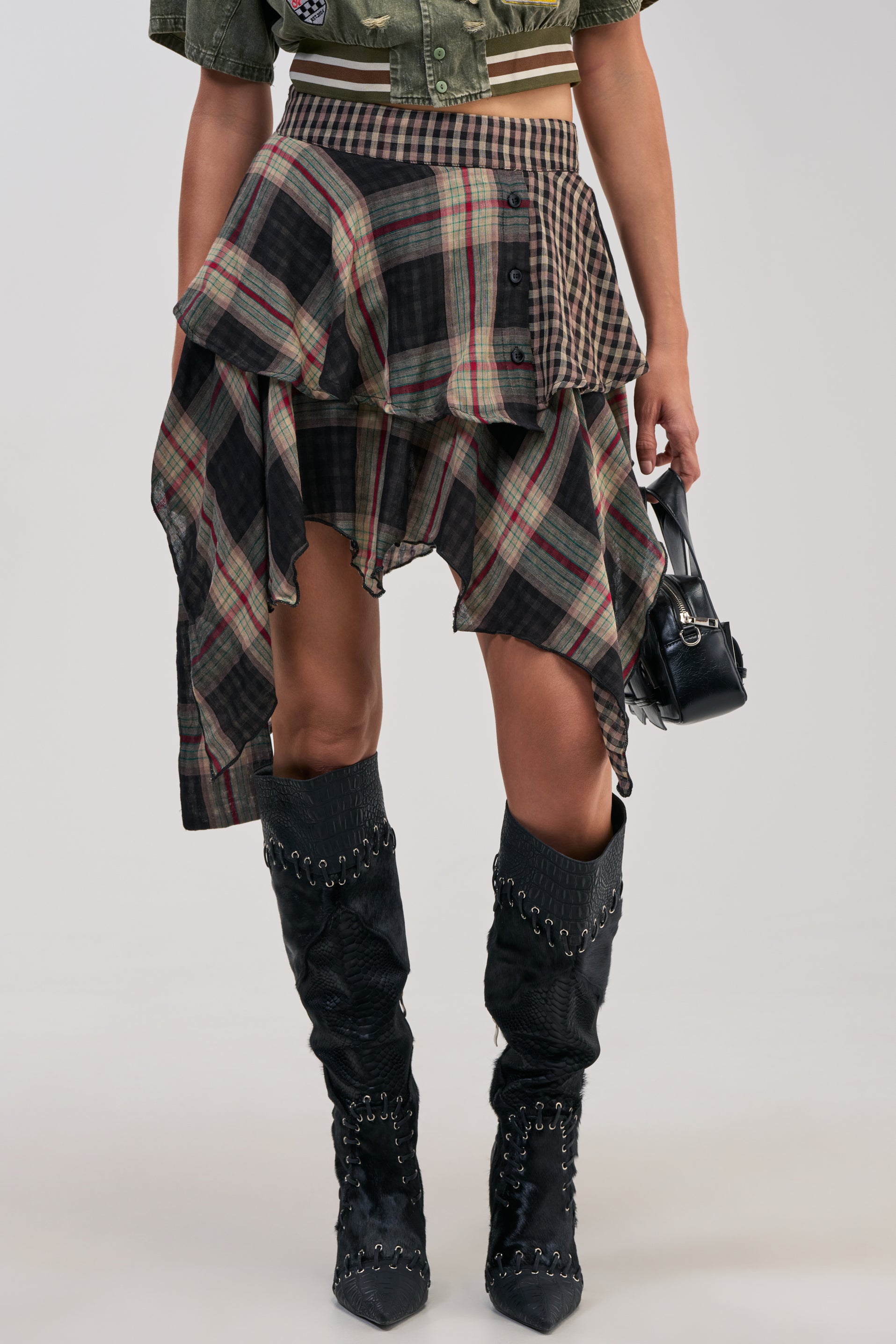 Wearing the MIXED EMOTIONS PLAID MINI SKIRT with its asymmetrical cut, this outfit pairs a cropped green shirt and black knee-high boots featuring chain details, accessorized with a small black handbag. The streetwear-inspired ensemble combines patterns and textures for a striking, bold look.