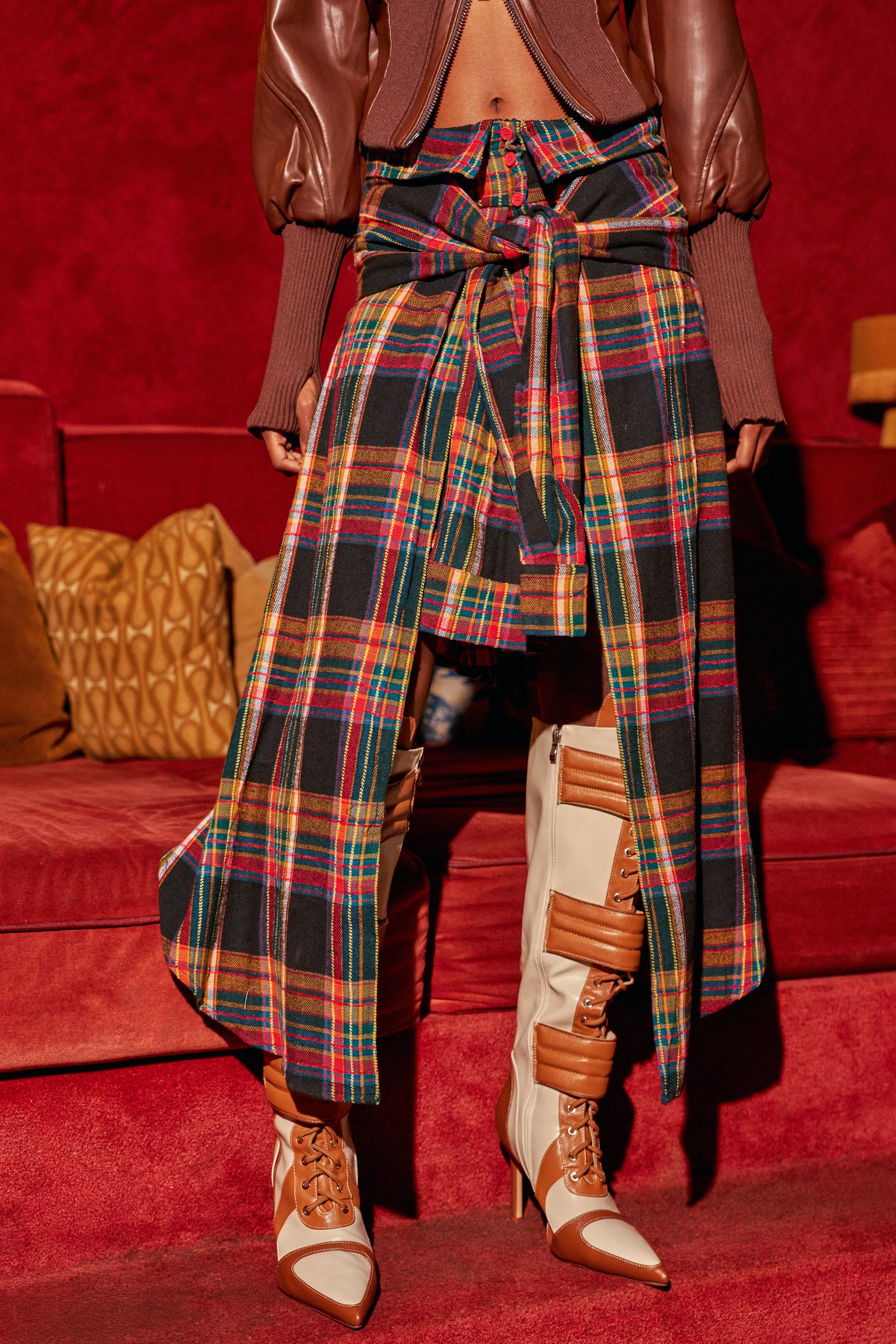 A person wears the PLAID FOR DAYS SKIRT, an asymmetrical plaid design with a tie-waist over a shorter skirt, paired with knee-high heeled boots featuring lace-up details and buckle straps.