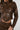 The person is wearing the UP TO IT LACE UP MOTO, a brown faux leather jacket with eyelet details and lace-up moto accents on the front and sleeves. The matching pants complete the cohesive ensemble, while their long, braided hair is partially visible.